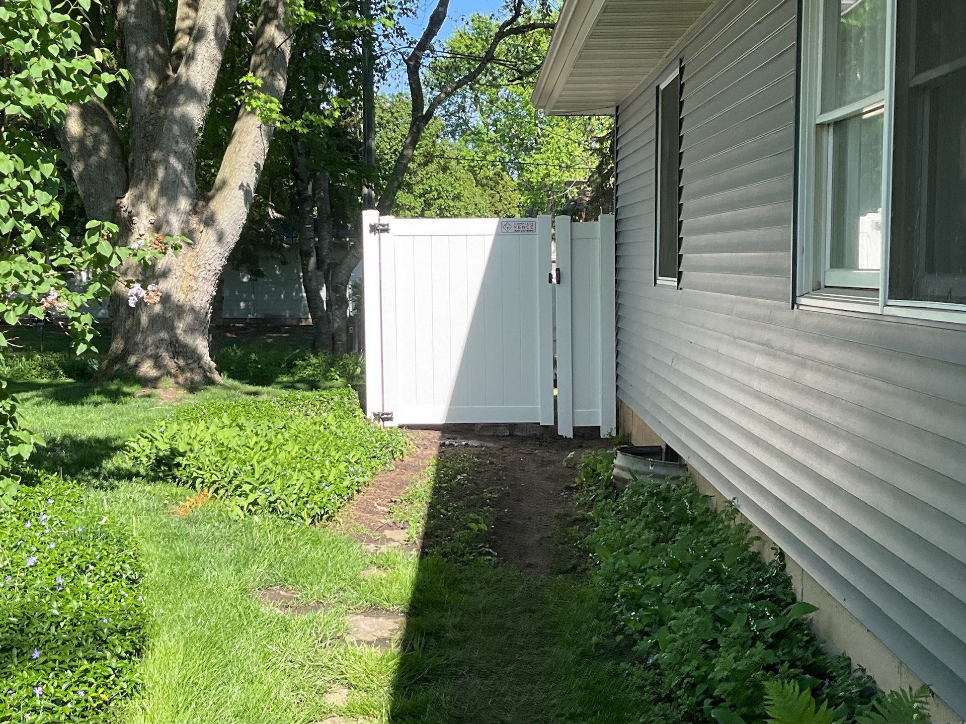 White fence next to a house with gray siding; a path in between green plants and grass.