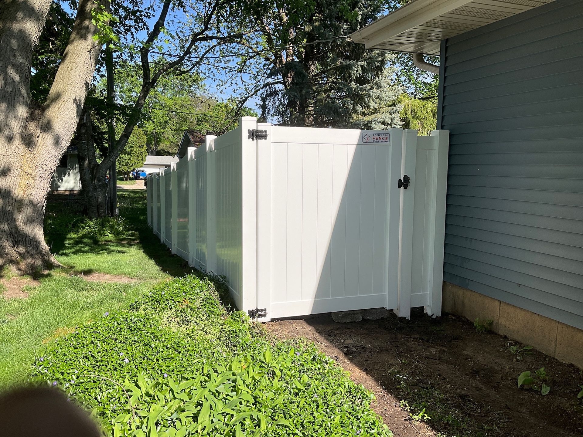 White vinyl fence with gate next to a light blue house. Green grass and trees are in the background.