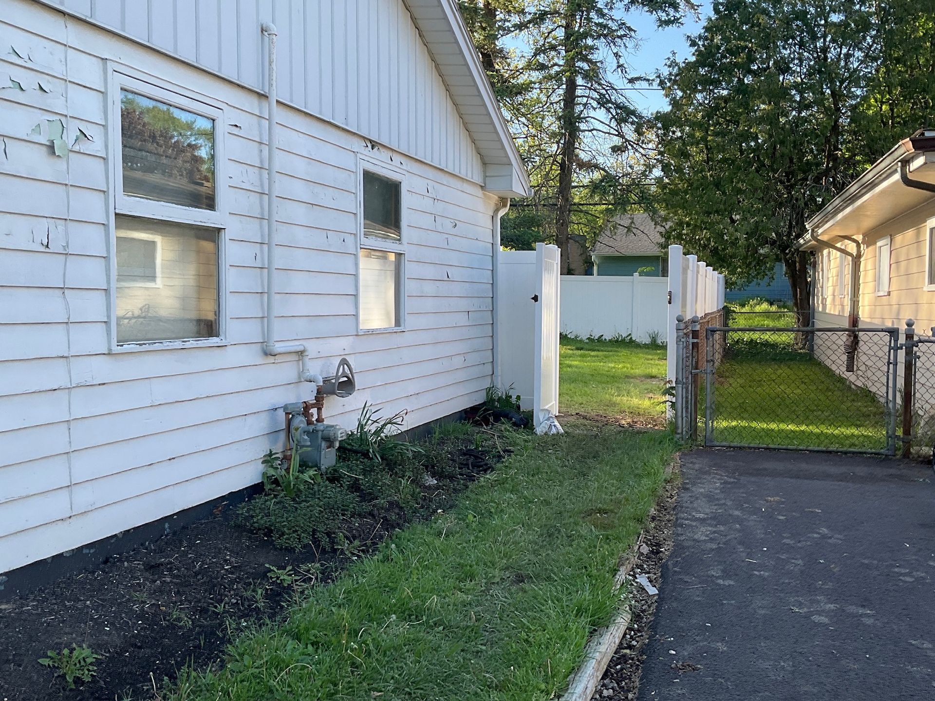 Side of a white house with a narrow strip of greenery, asphalt path, and a white fence leading to a backyard.