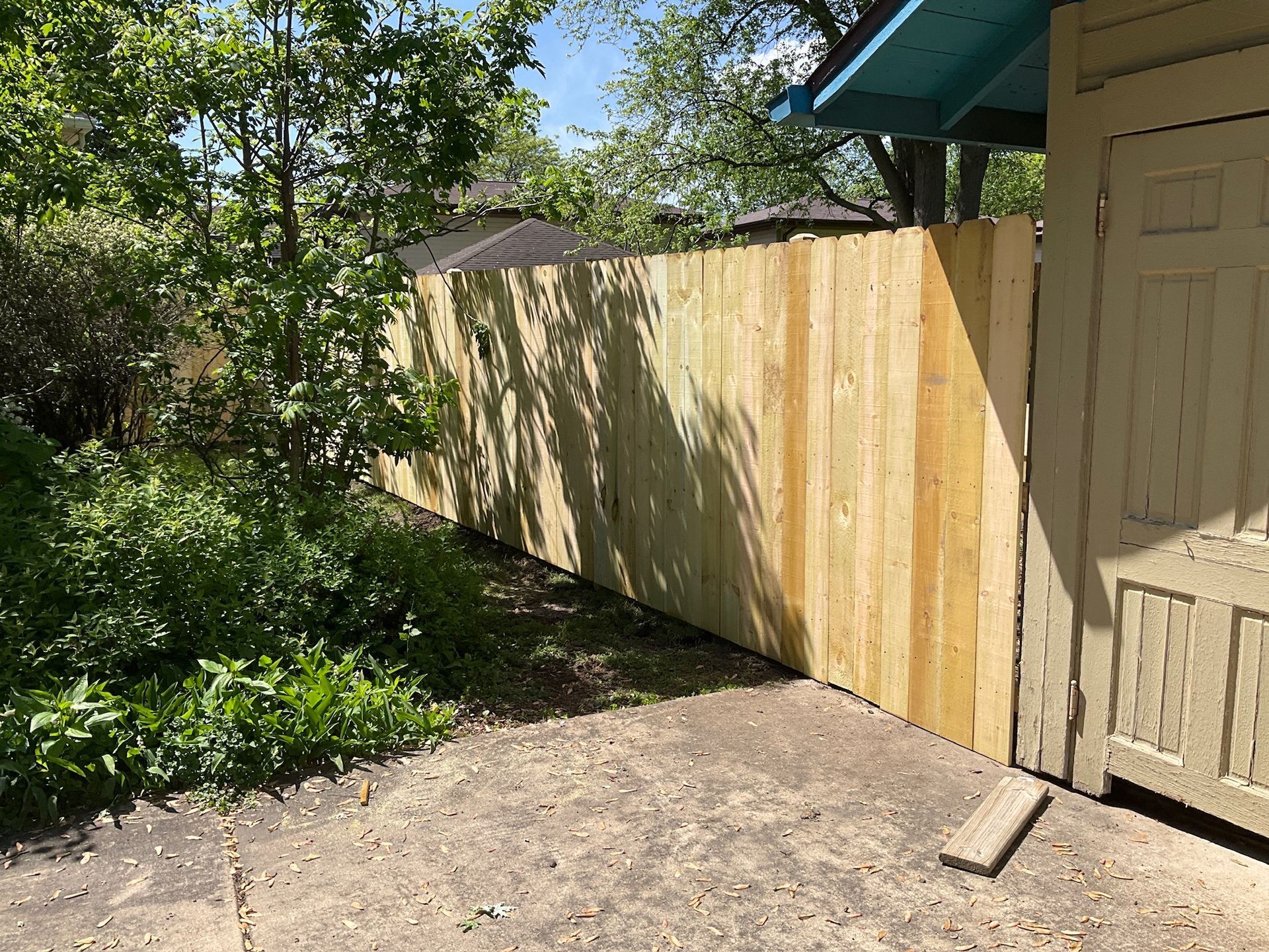 Wooden fence next to a beige building, near trees and concrete ground.