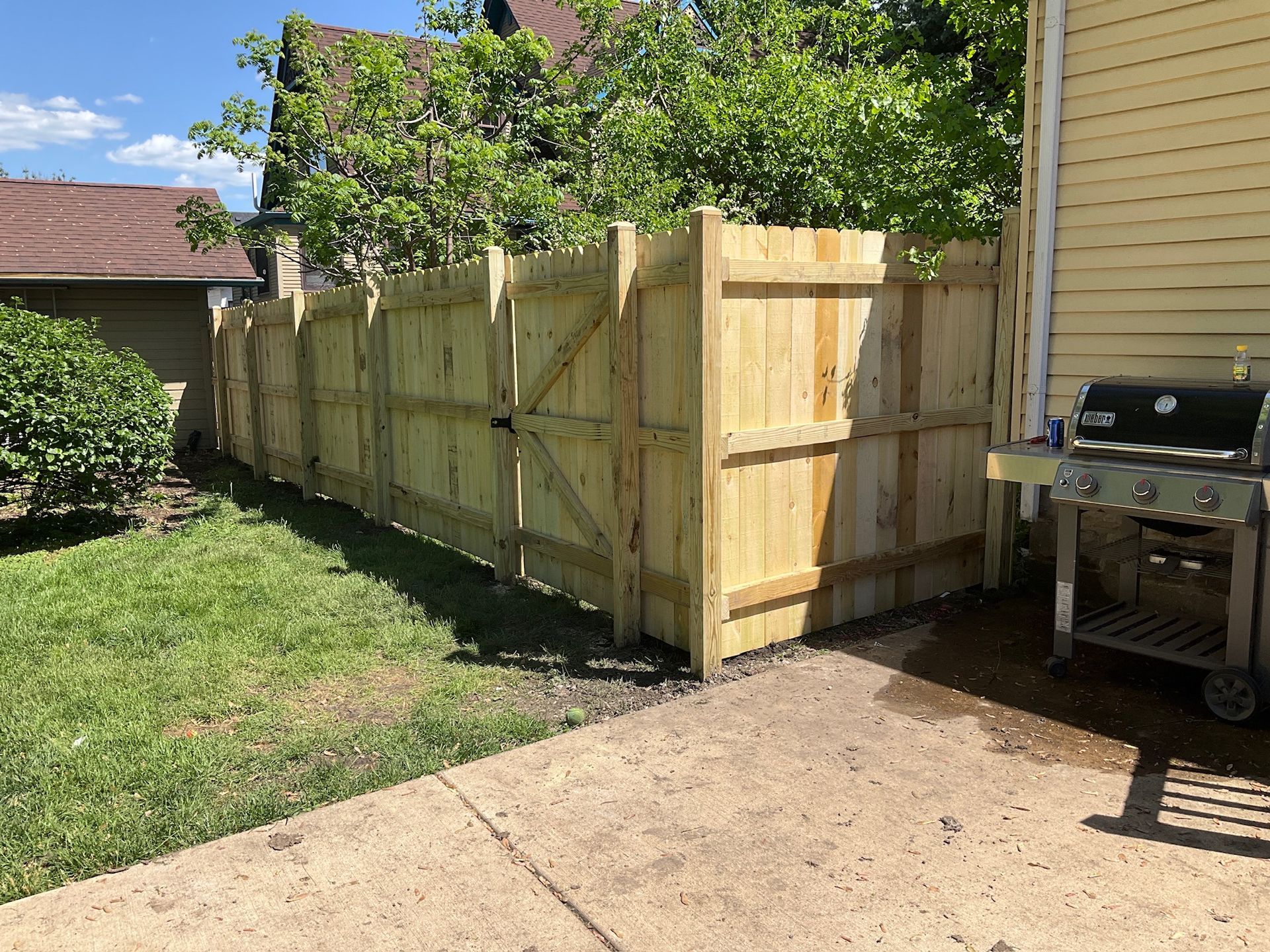 Wooden fence in backyard next to a patio and grill, with green grass and foliage.