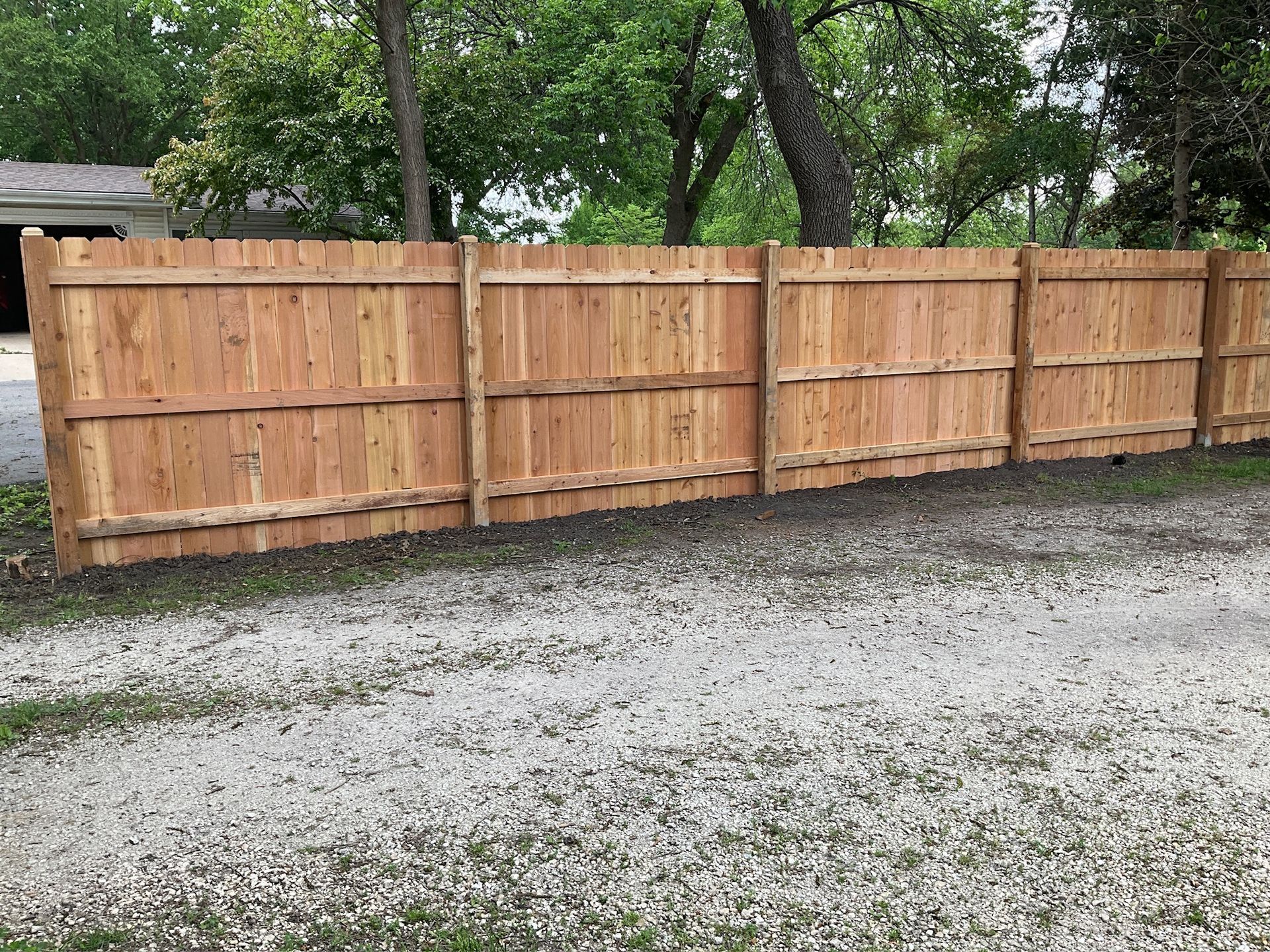 Wooden fence in front of a gravel driveway, trees in the background, daytime.