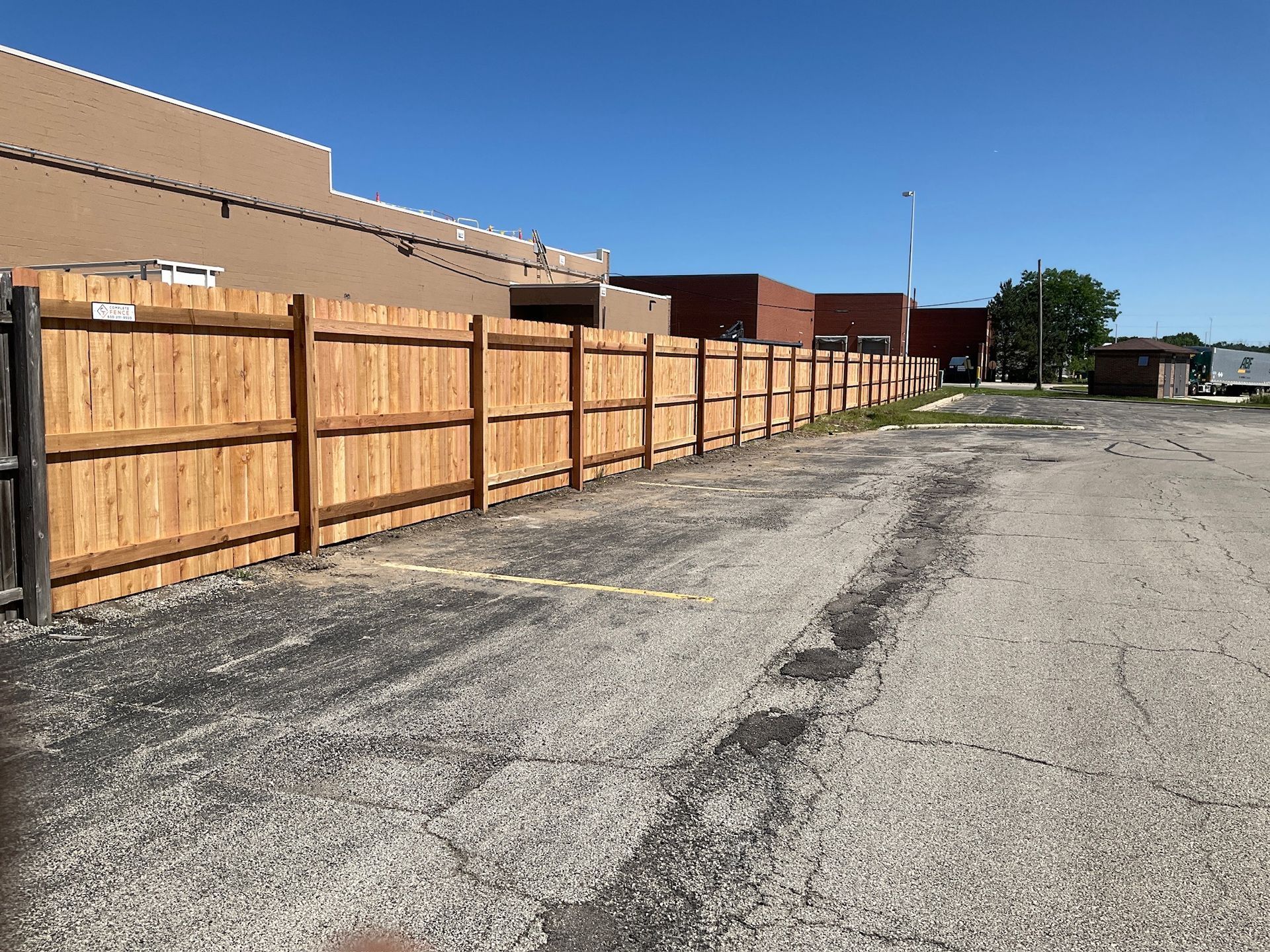 Wooden fence alongside an asphalt parking lot with buildings in the background under a blue sky.