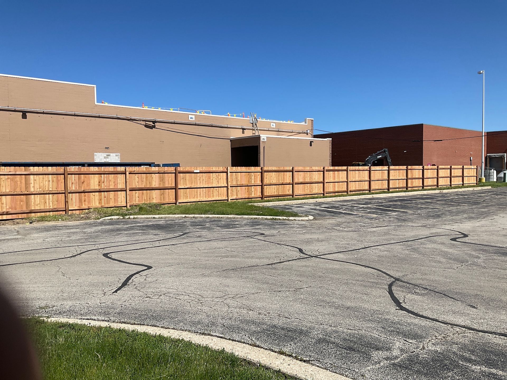 Wooden fence in front of a tan and red building. Asphalt parking lot in the foreground, clear blue sky.
