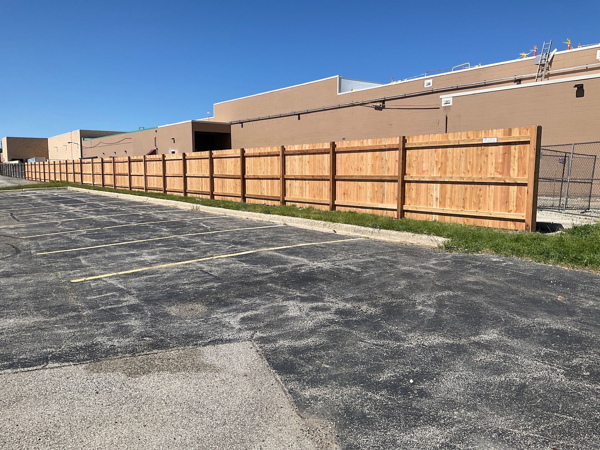 Wooden fence along a parking lot next to a tan building on a sunny day.