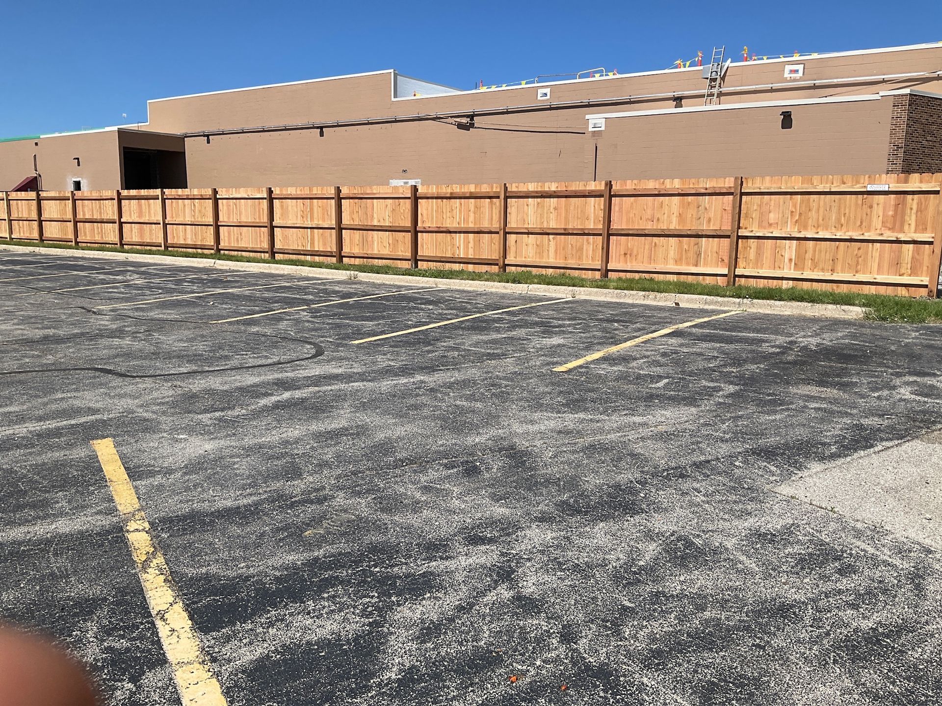 Wooden fence beside an empty parking lot, tan building in the background. Bright blue sky.