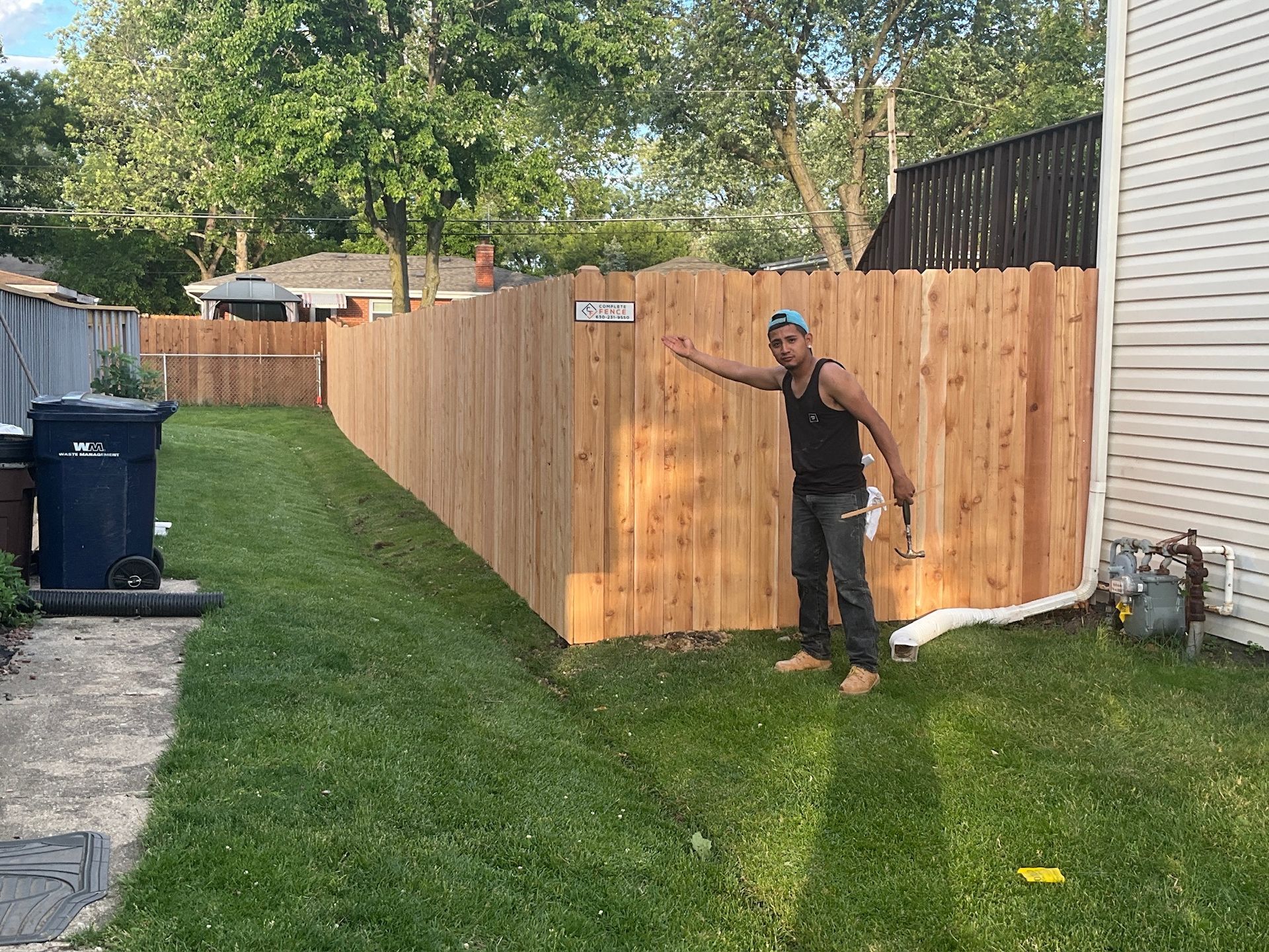 Person points at a new wooden fence in a yard with green grass. A trash can is on the left.