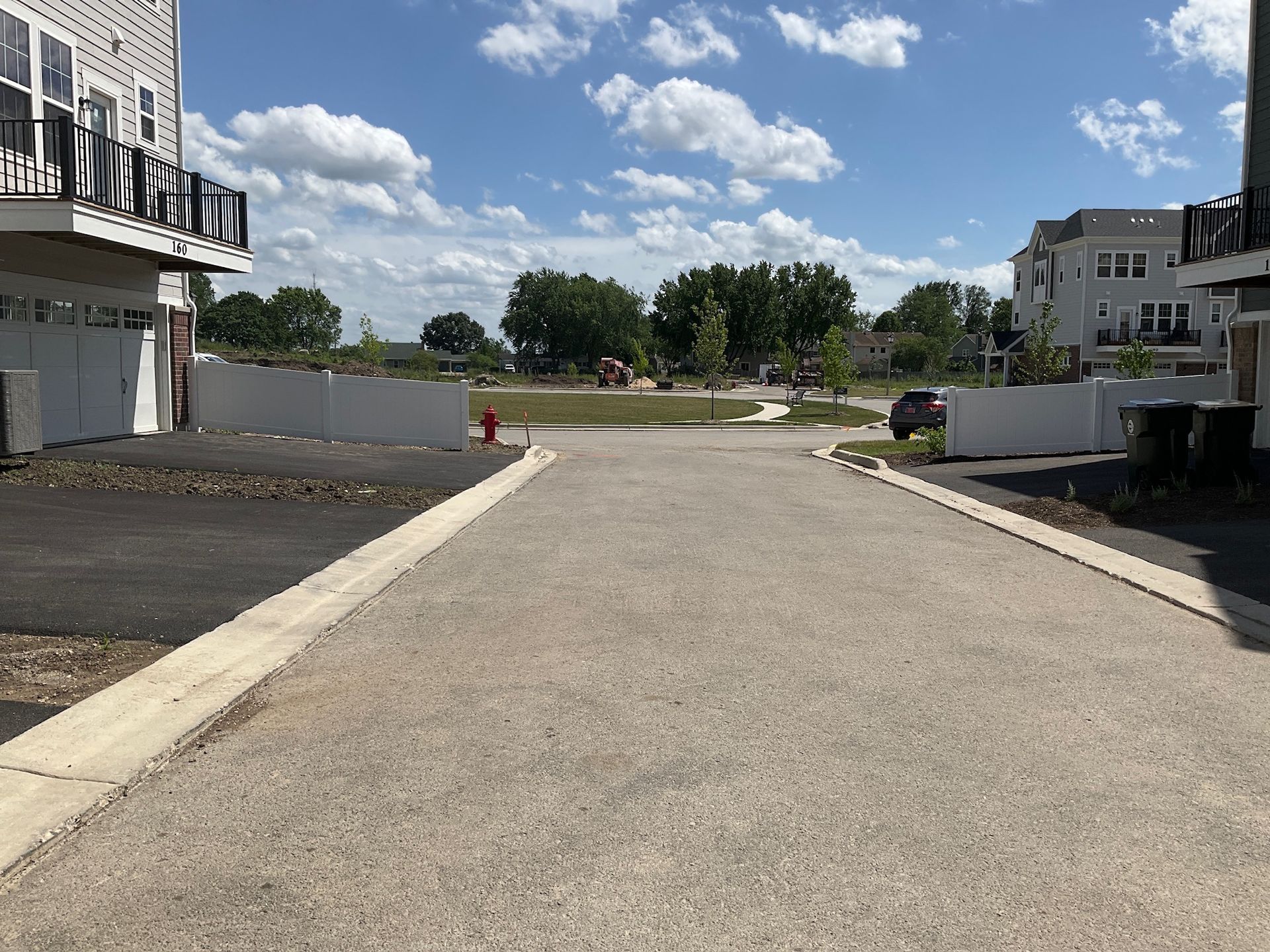 Paved road in a residential area, leading toward a park. White buildings, blue sky with clouds.