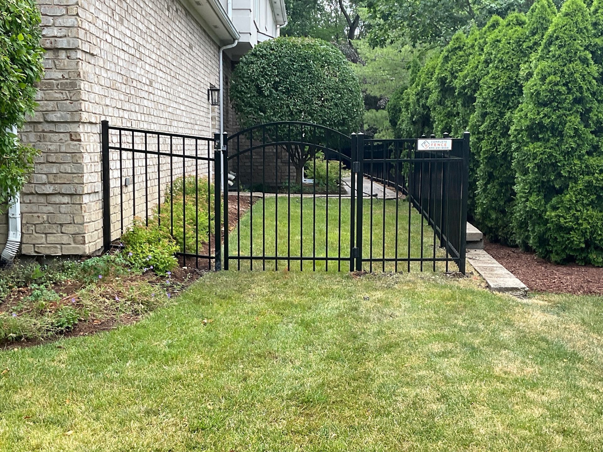 Black metal fence surrounding a small, grassy yard next to a brick building and bushes.