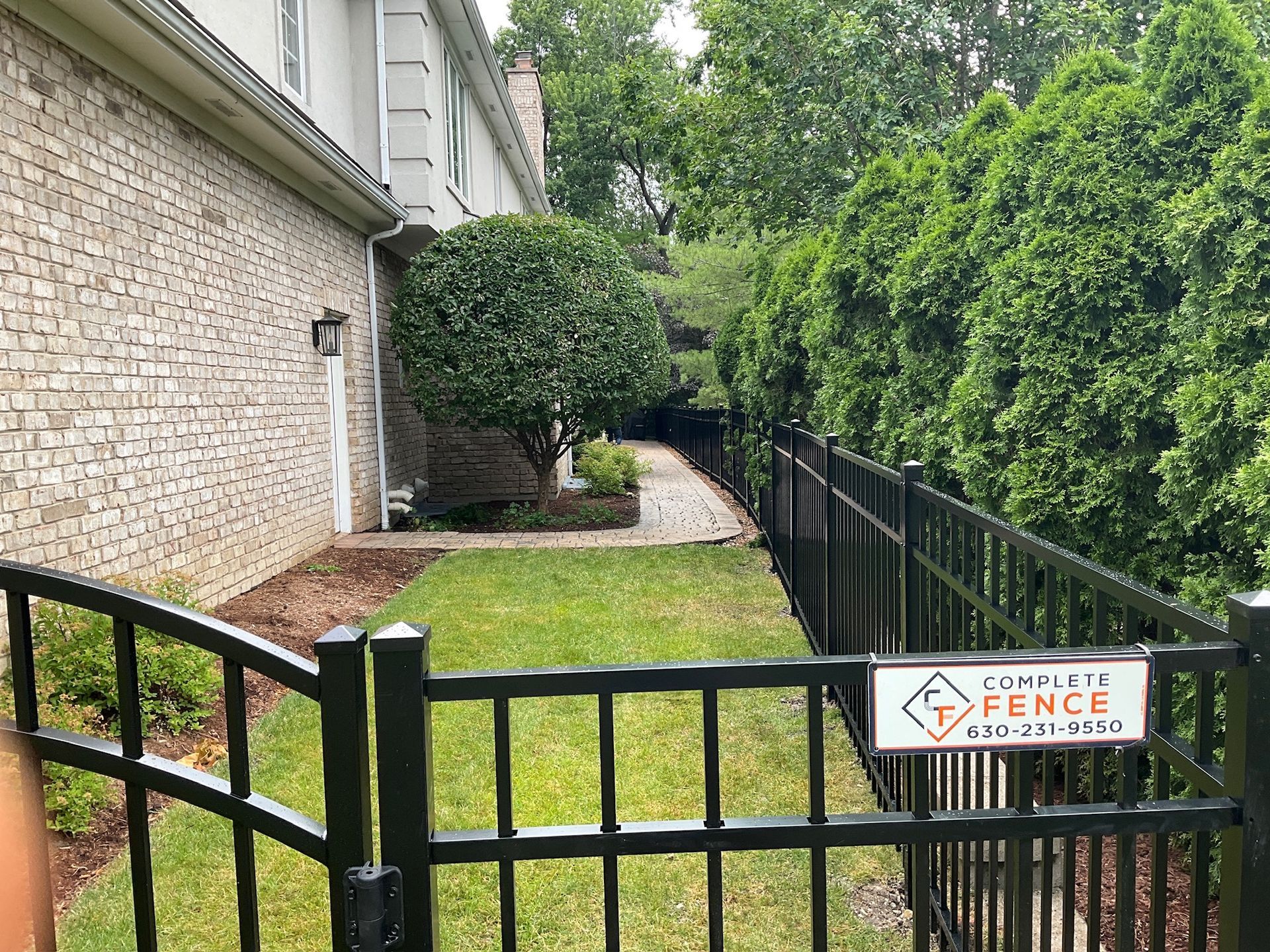 Black metal fence along a brick house and a green yard with a Complete Fence sign.