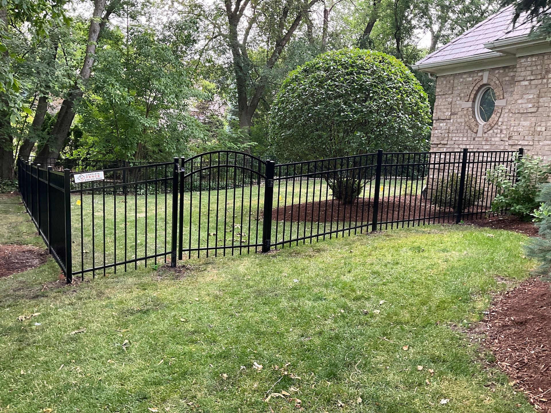 Black metal fence enclosing a grassy yard with trees and a brick building in the background.