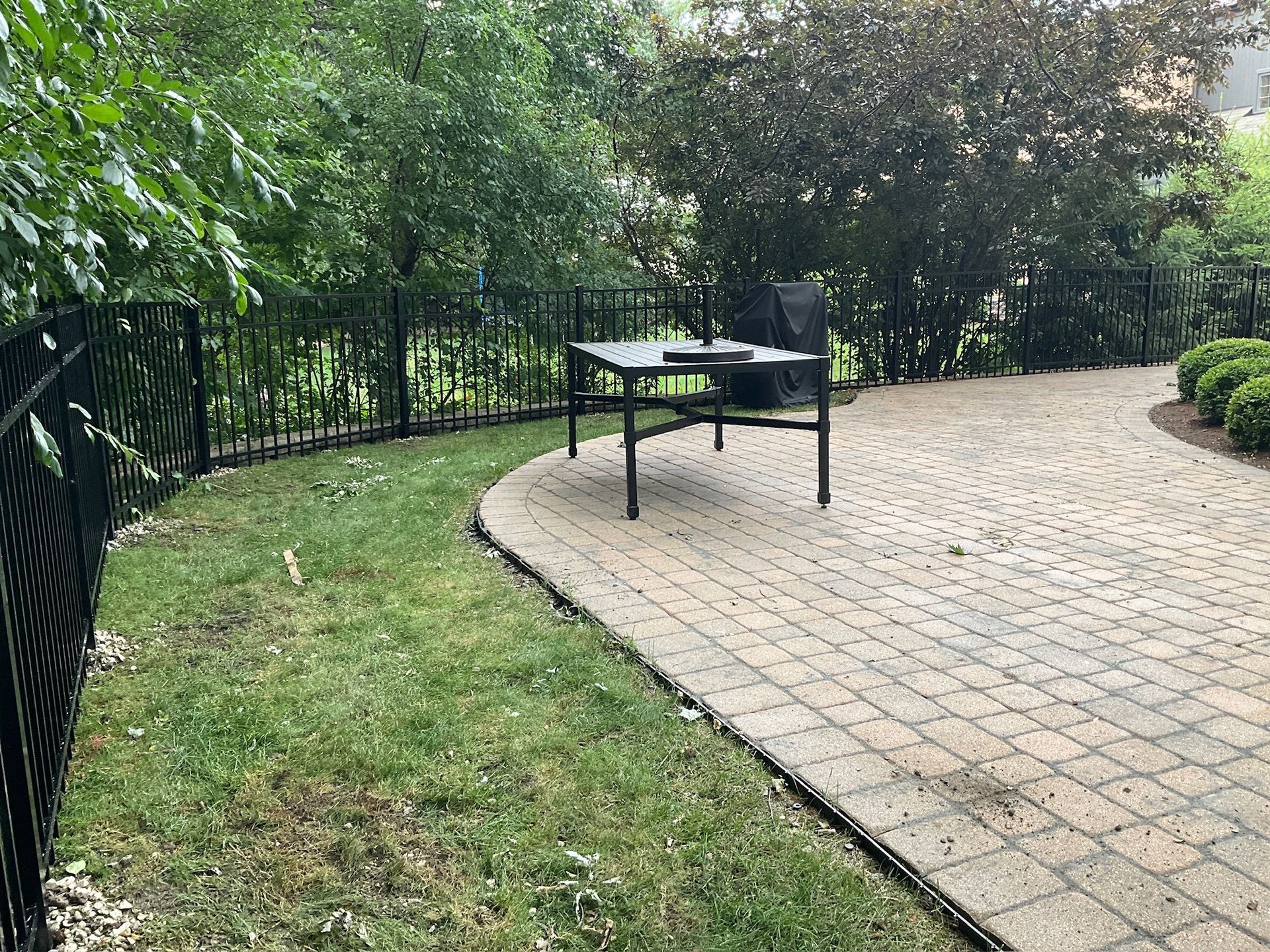 Patio with brick pavers, table, chairs, and black metal fence in a yard with trees and grass.