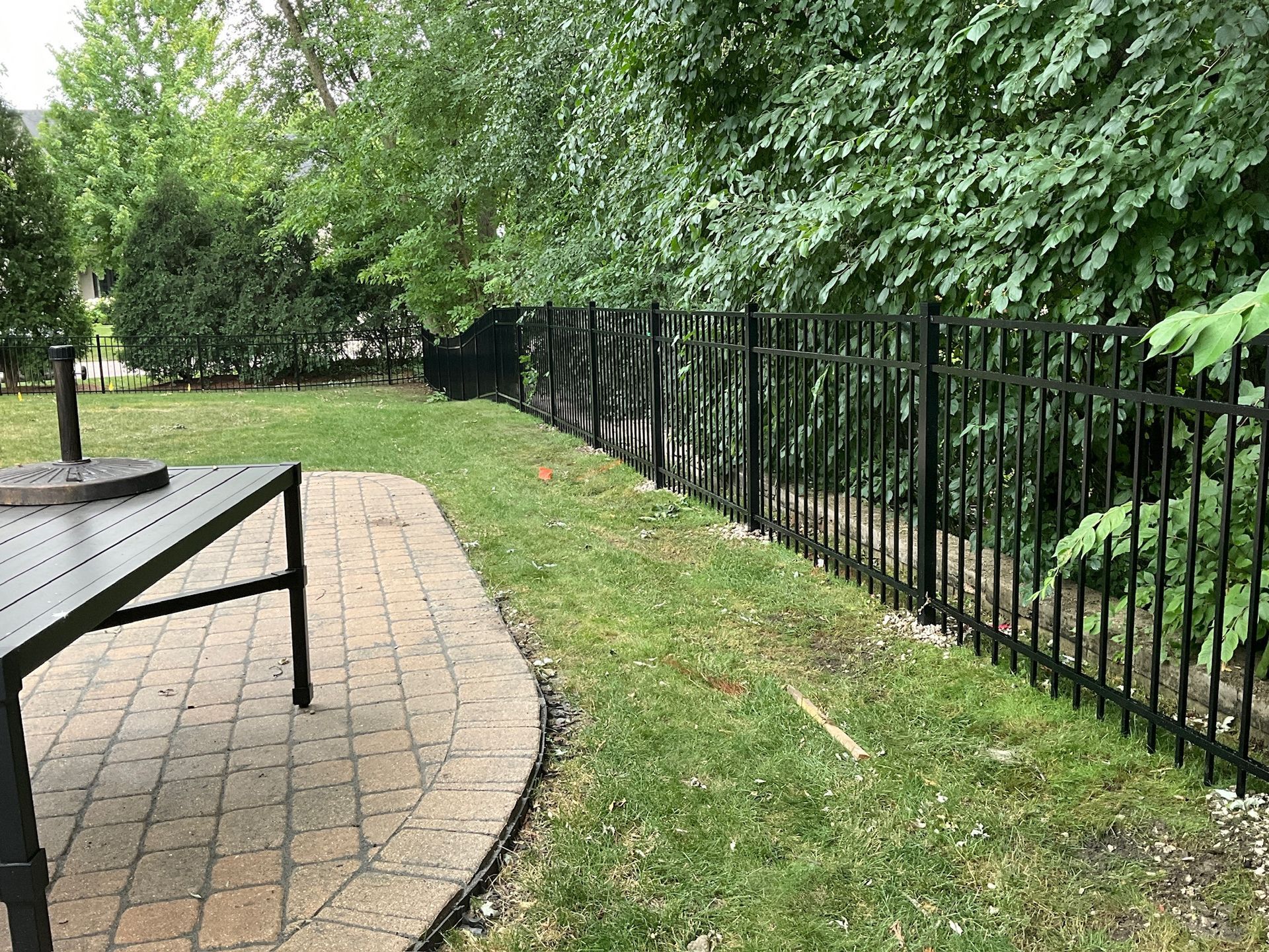 Black metal fence bordering a green lawn and trees, with a stone patio and black table in the foreground.