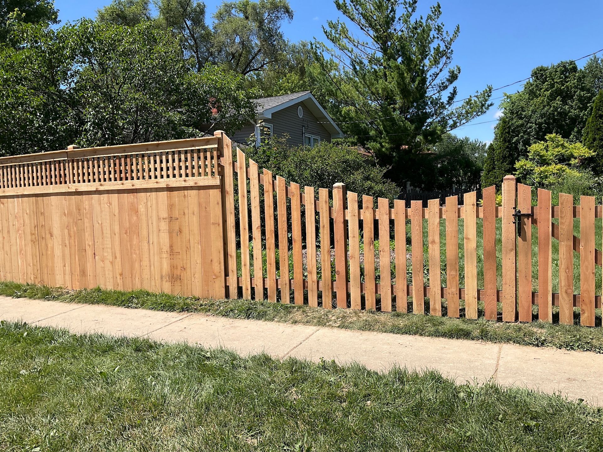 Wooden fence with a scalloped edge next to a sidewalk and green grass, with trees and a house in the background.
