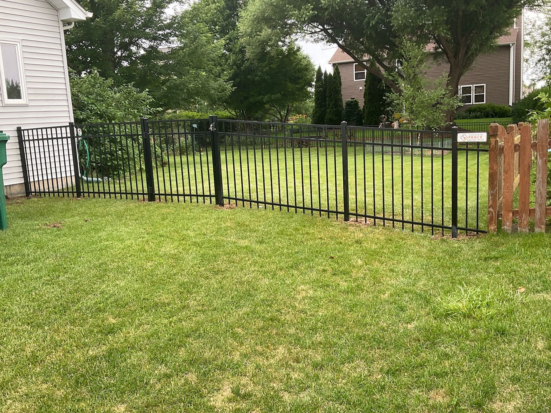 Black metal fence encloses a green lawn, next to a house and other trees.