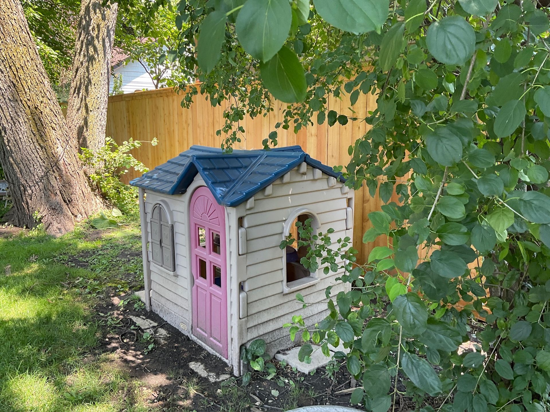 Playhouse with pink door and blue roof in a backyard, surrounded by greenery and a wooden fence.