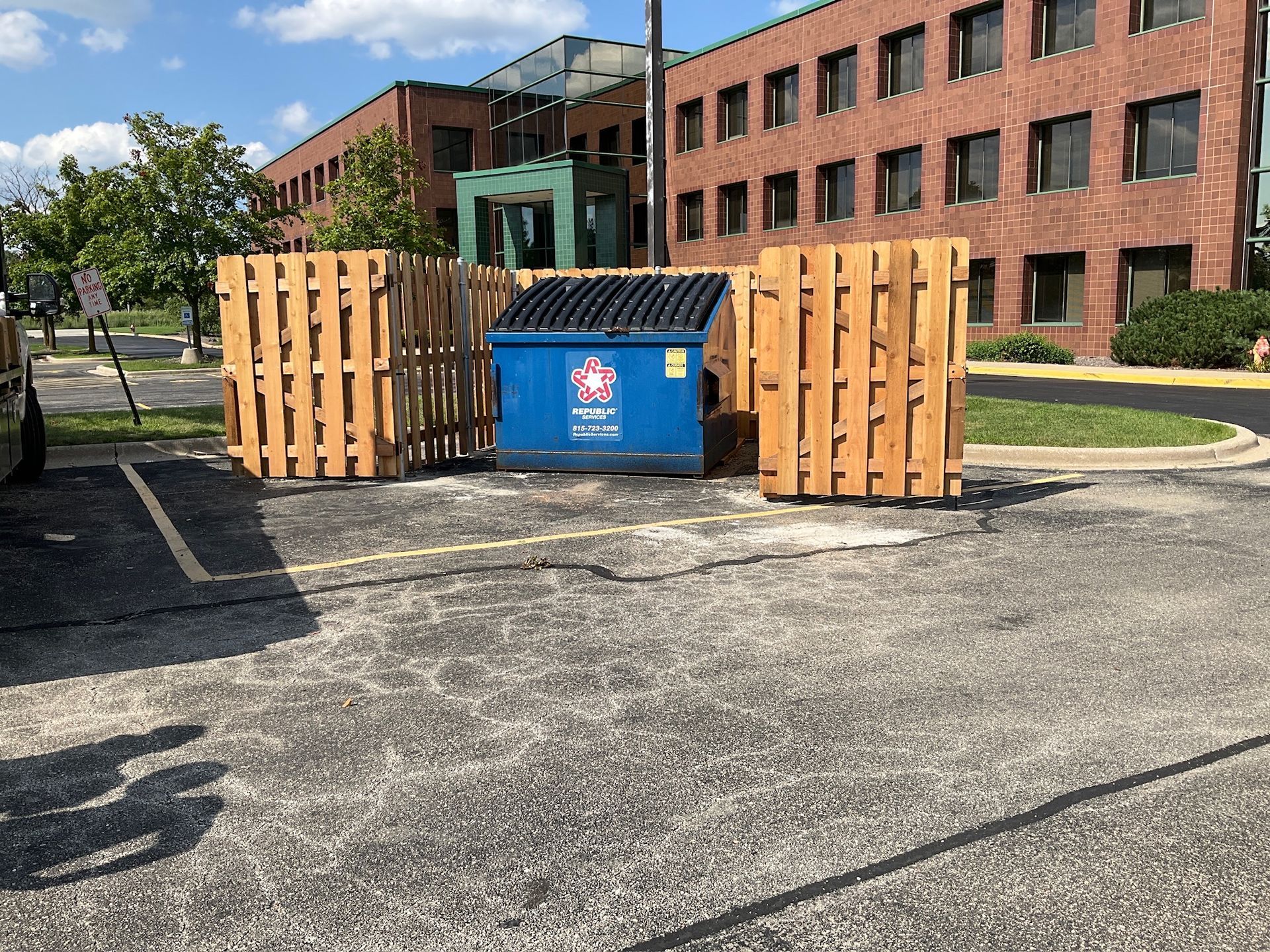 Blue dumpster enclosed by wooden fence in a parking lot, near a brick building.