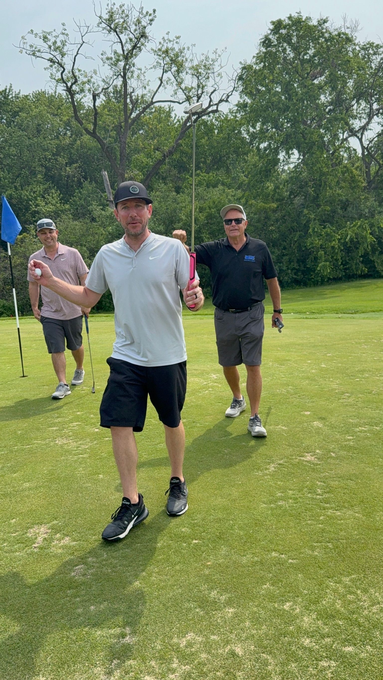 Three men on a golf course celebrate a shot. The central man pumps his fist, holding a flagstick.