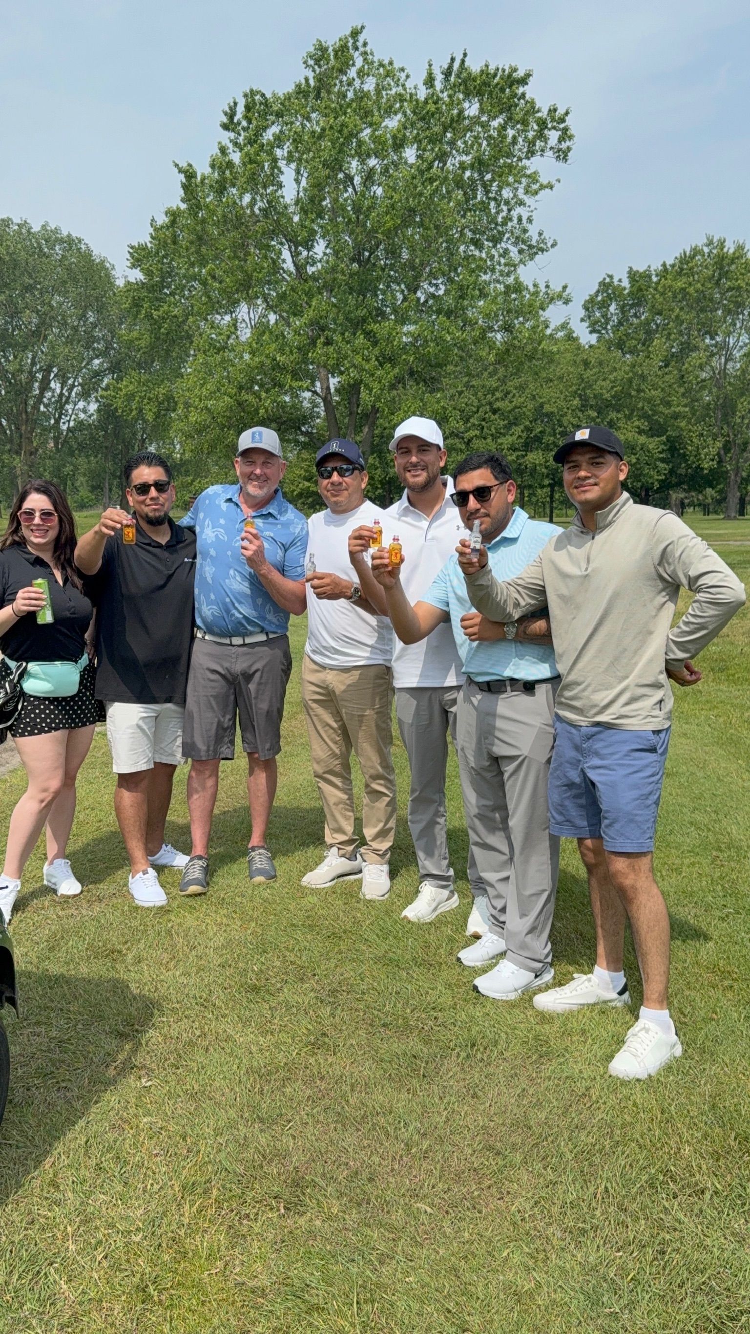Group of people on a golf course holding donuts, smiling. Sunny day, trees in background, wearing casual attire.