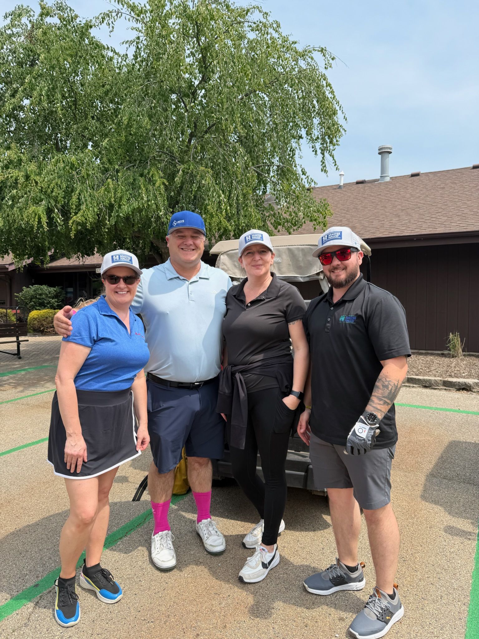 Four people in golf attire pose outdoors near a building. All wear hats with a logo.