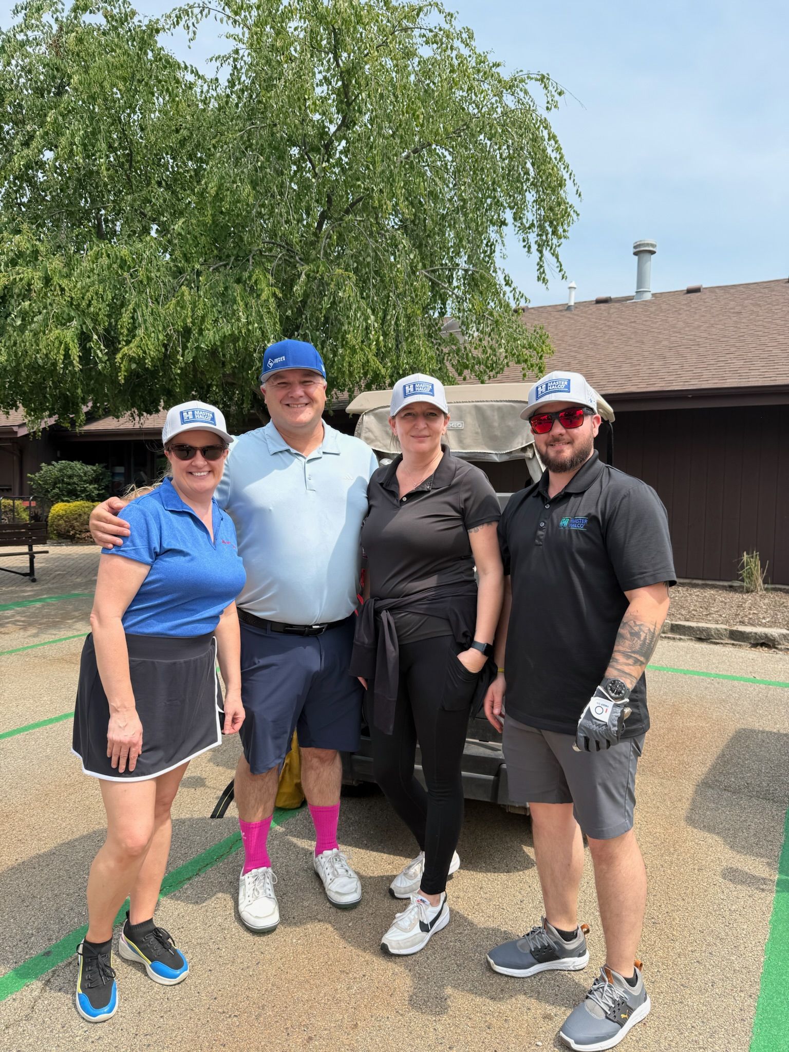 Four people pose outdoors, near a golf cart. Two women and two men wearing golf attire. Blue and black clothing. Sunlight.