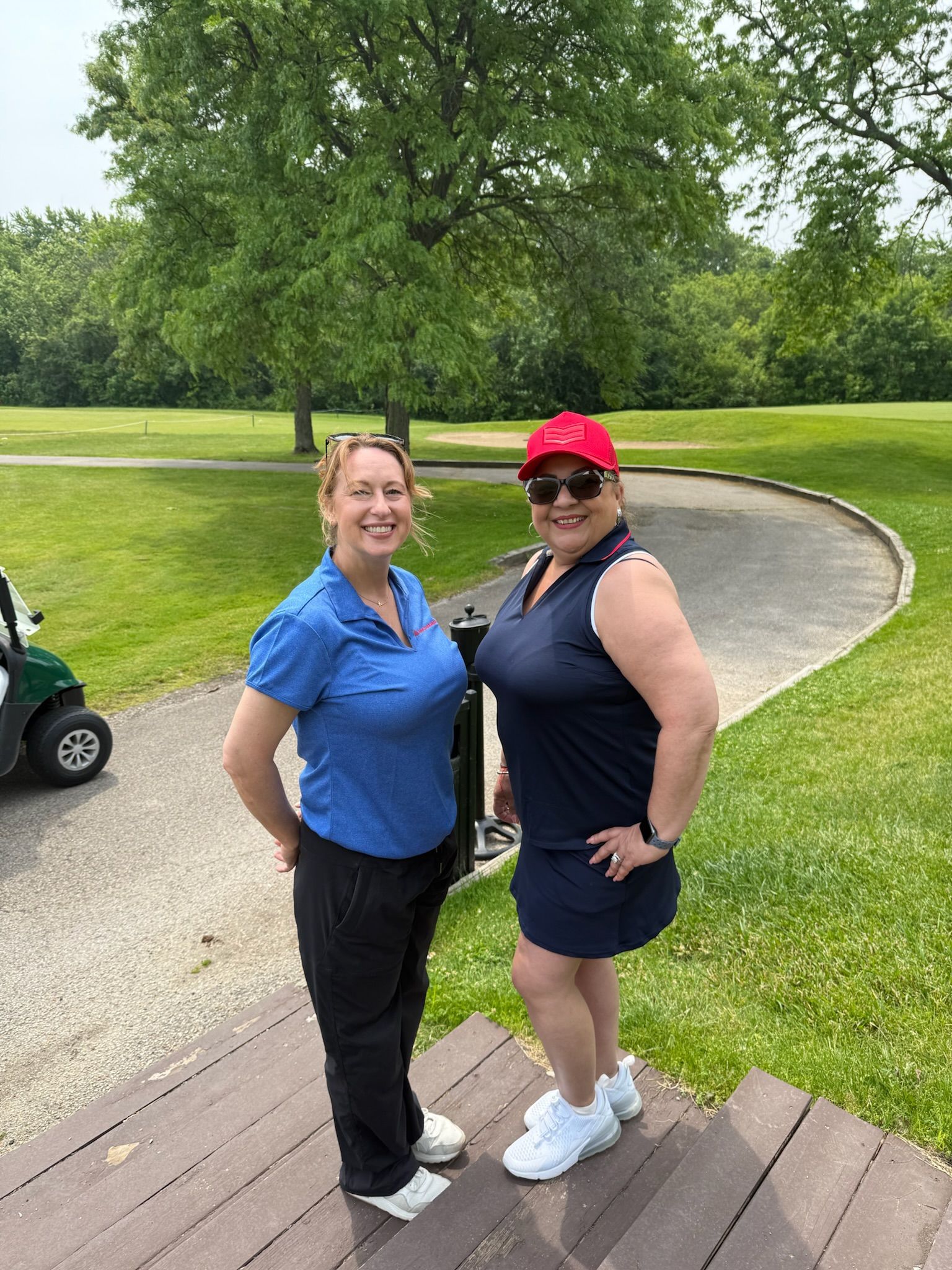 Two women on a golf course. One wears blue, the other navy with a red hat. They smile, posing near a cart.