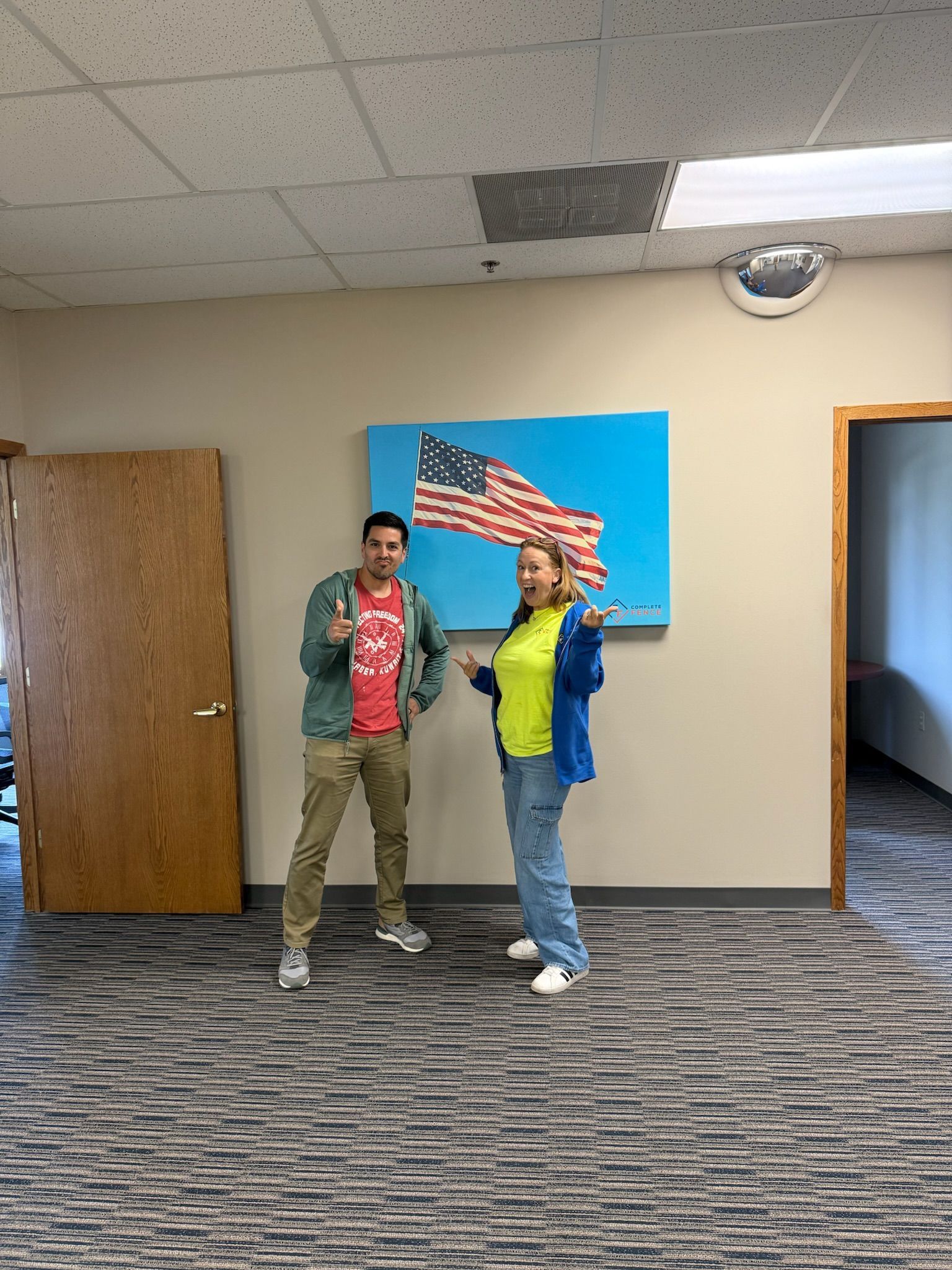 Two people pose in front of an American flag painting; man gives thumbs up, woman gestures with hands.