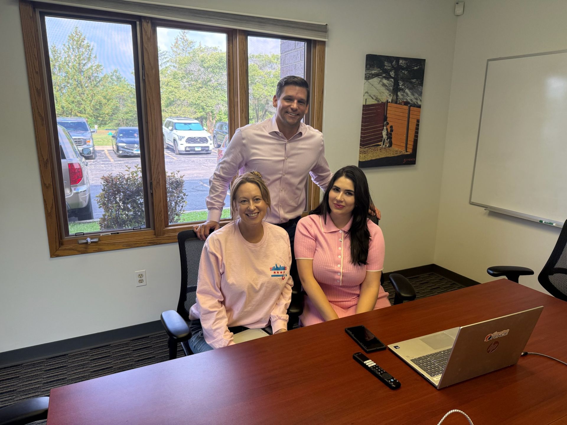 Three people in a brightly lit office; all wearing pink. Two women sit at a table, one man stands behind them.