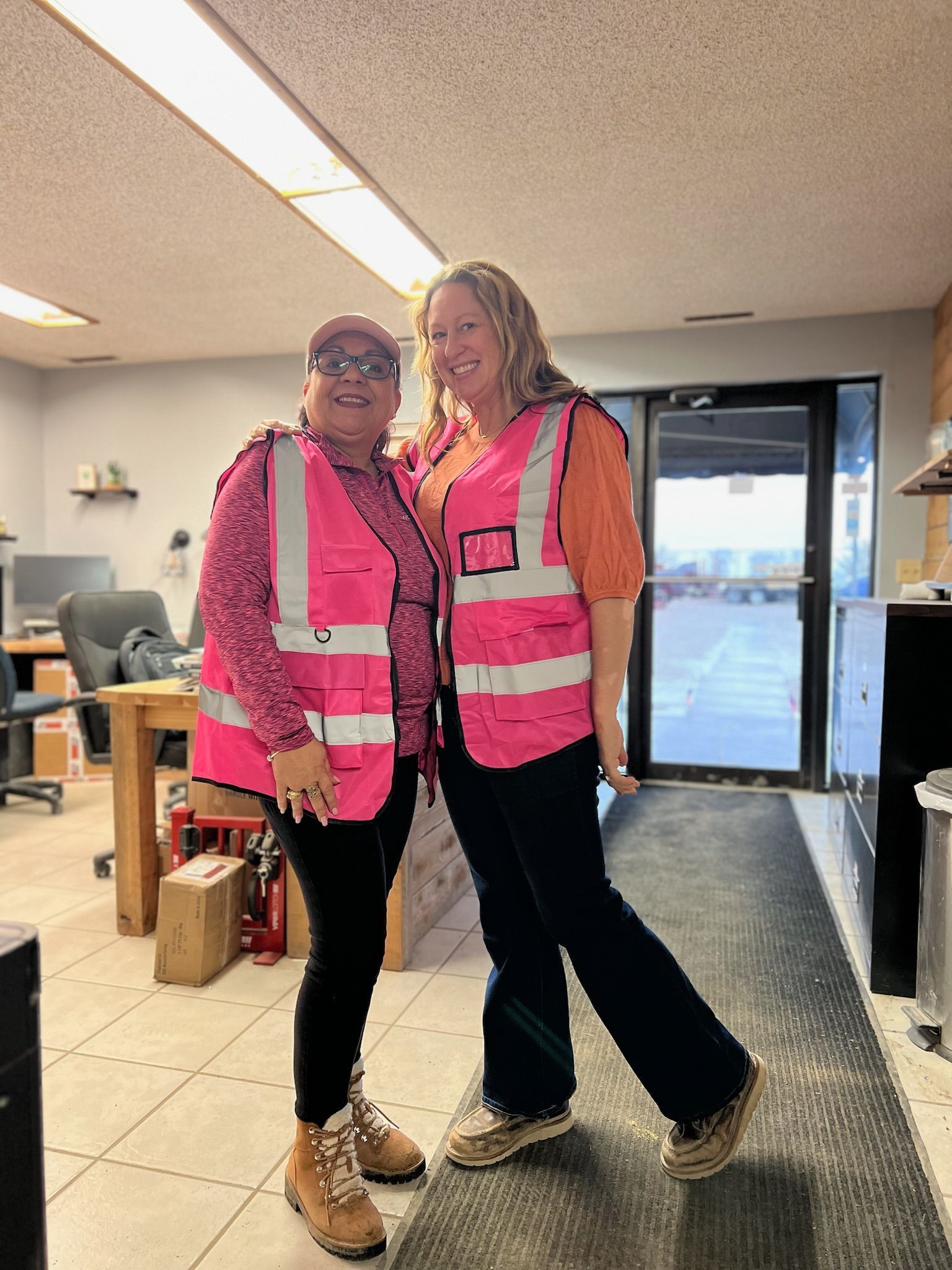 Two women in pink safety vests smile in an office; one poses with a leg extended.