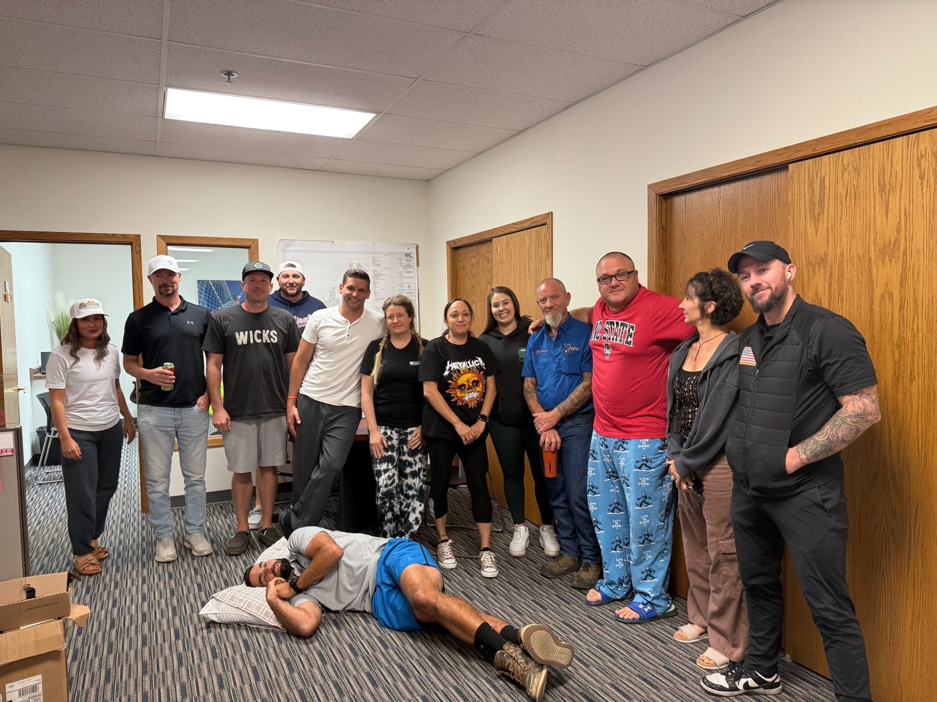 Group of people in an office. One person lies on the floor; others stand smiling.