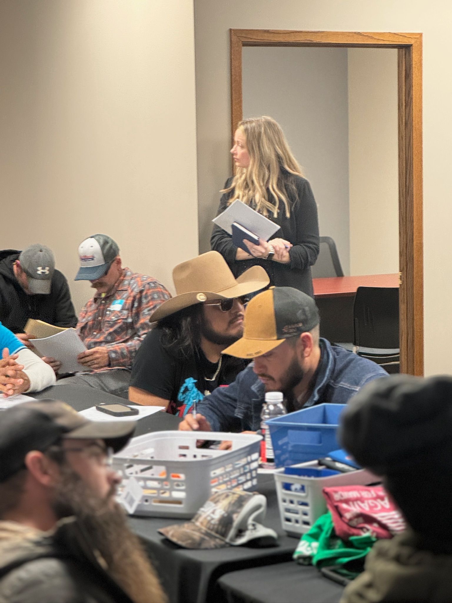 A woman stands in a doorway, holding paperwork, as a group of people sit at tables; a meeting or workshop in a room.