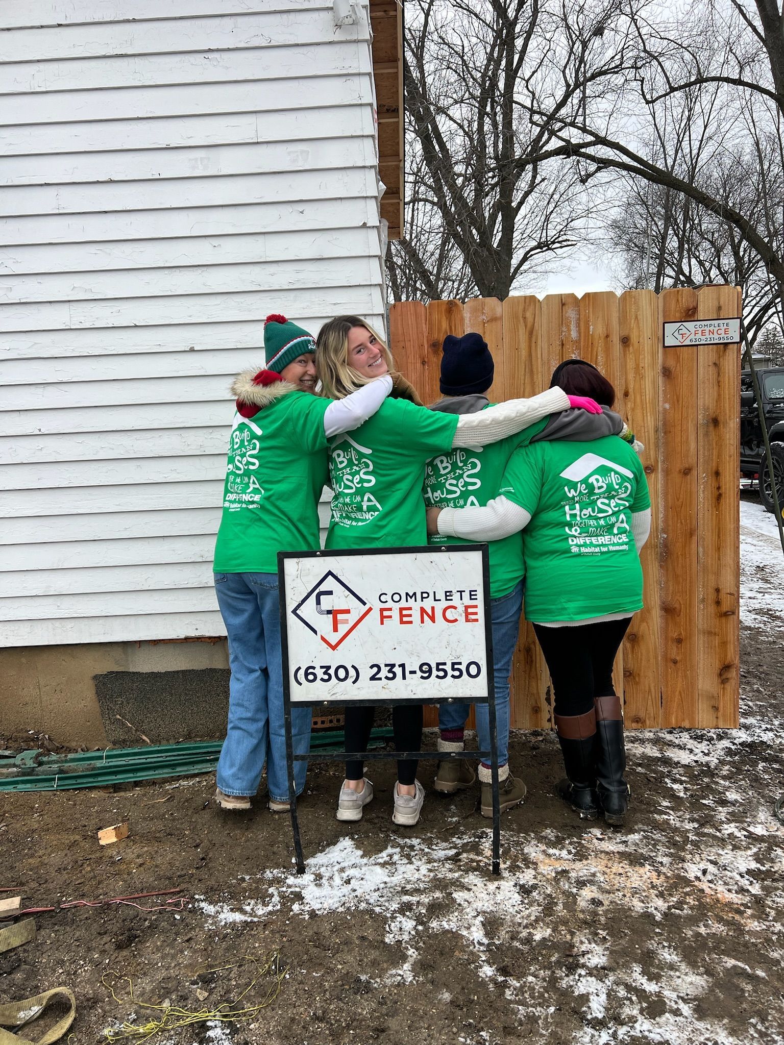 Four people in green shirts hug next to a wooden fence and sign. White building in the background.