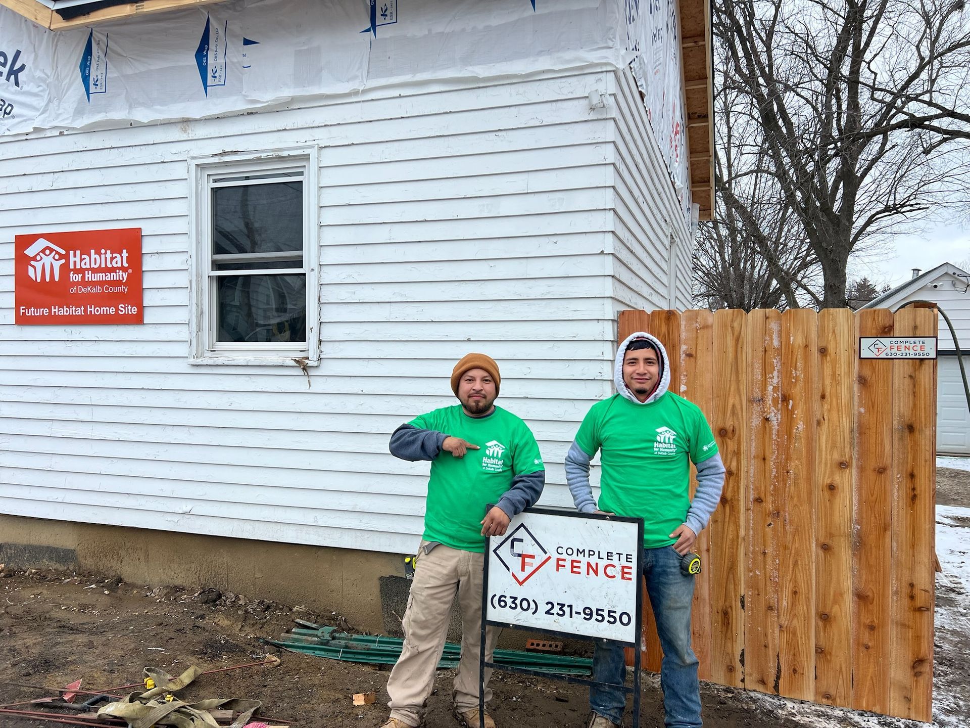Two people in green shirts stand next to a fence and house with a Habitat for Humanity sign.