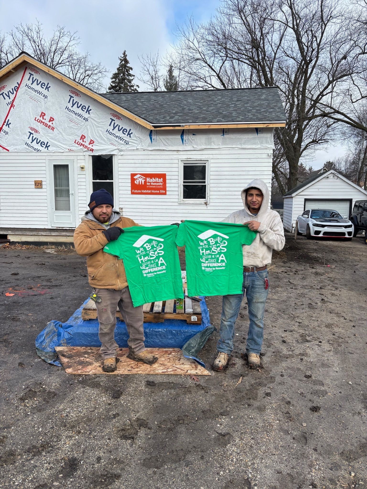 Two men holding green shirts with a house logo, standing in front of a house under construction on a cloudy day.