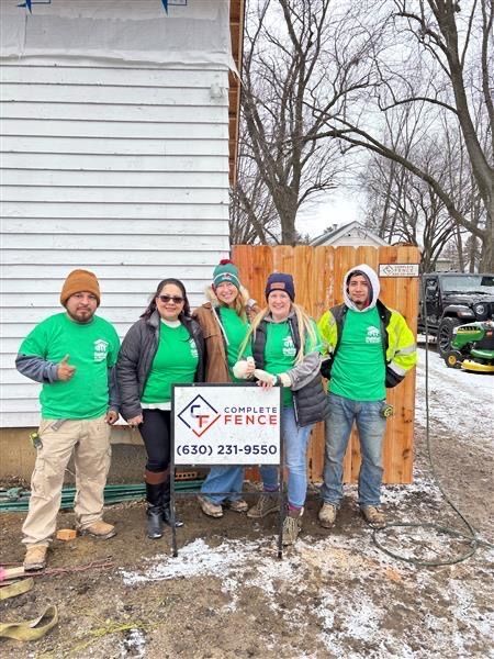 Five people in green shirts stand next to a fence and house. A sign displays a phone number.