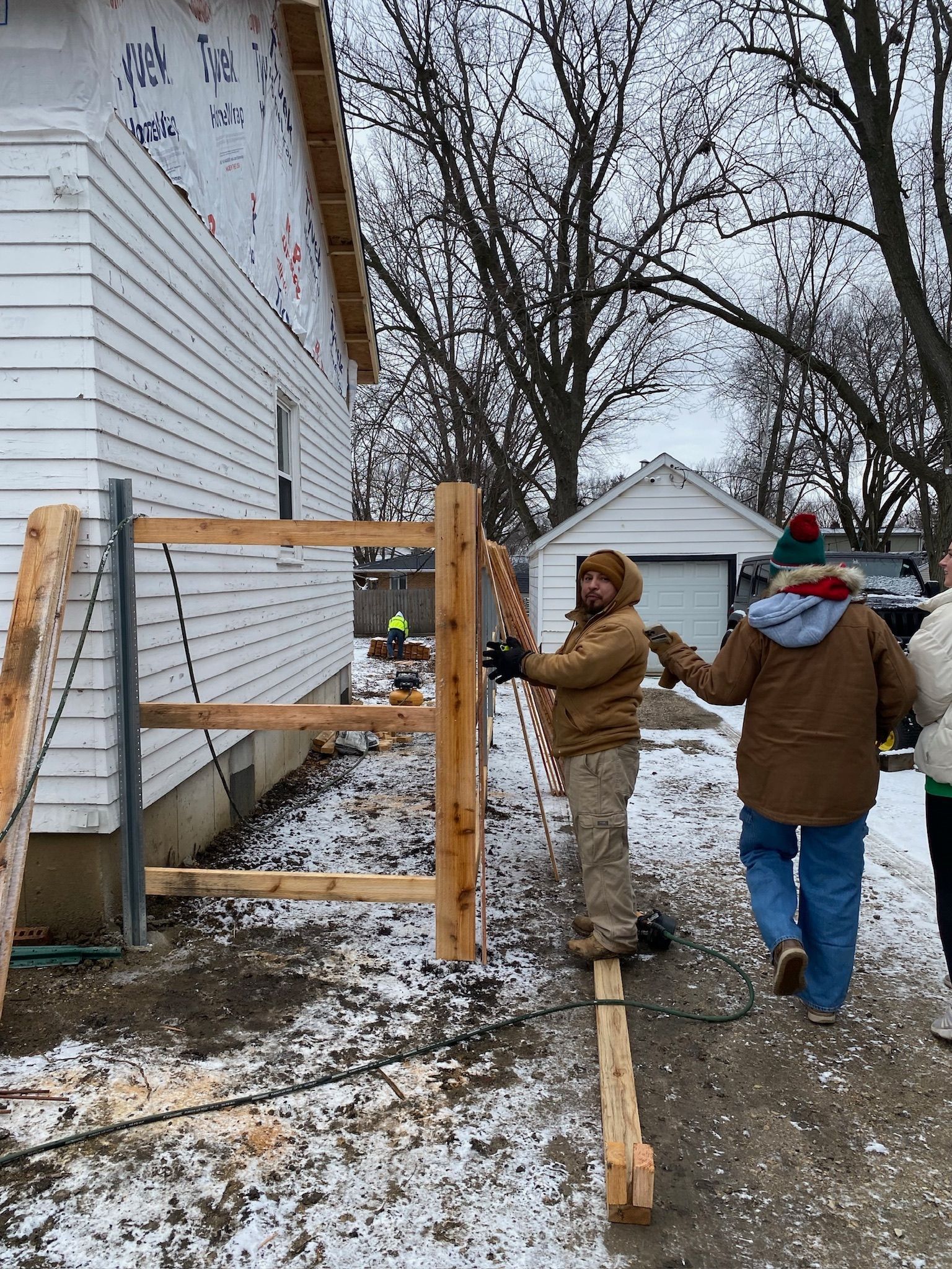 Two people carrying a wooden fence post in a snowy outdoor setting next to a building.