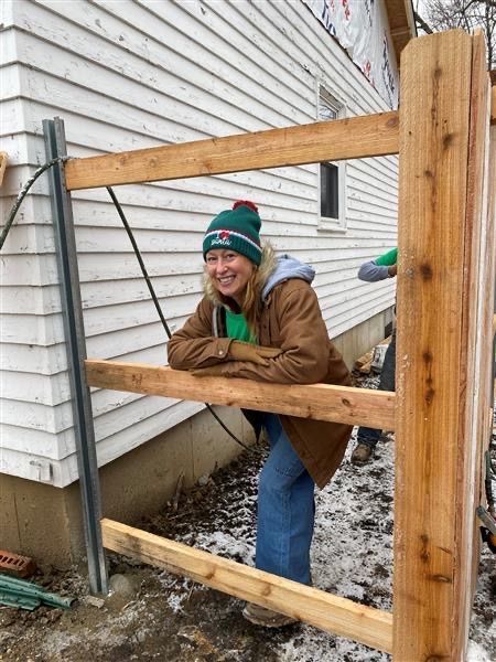 Woman smiles, leaning on a partially built wooden fence. Building in background, snowy ground.