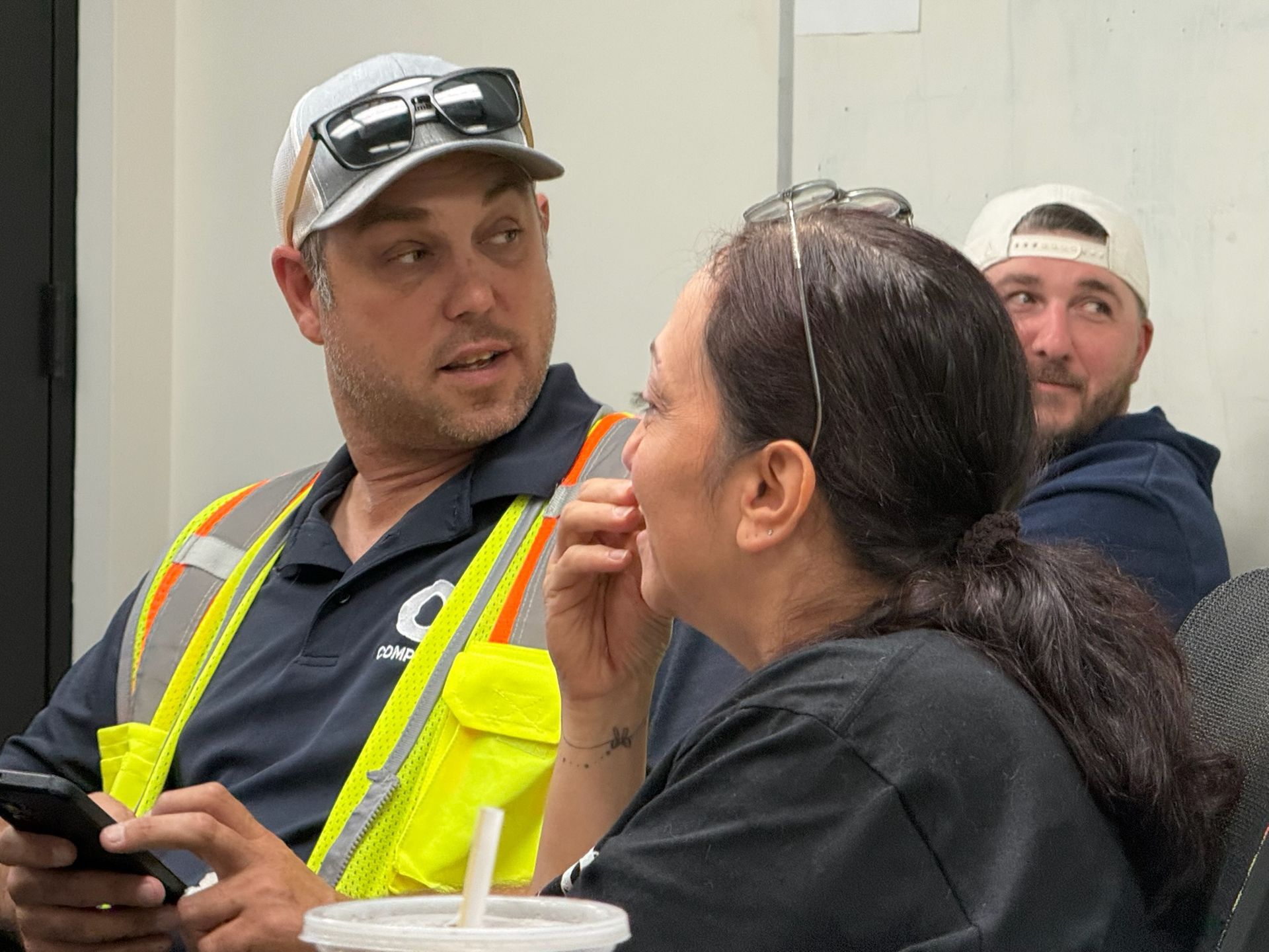 Man in safety vest talking to woman, both seated, another man in background.