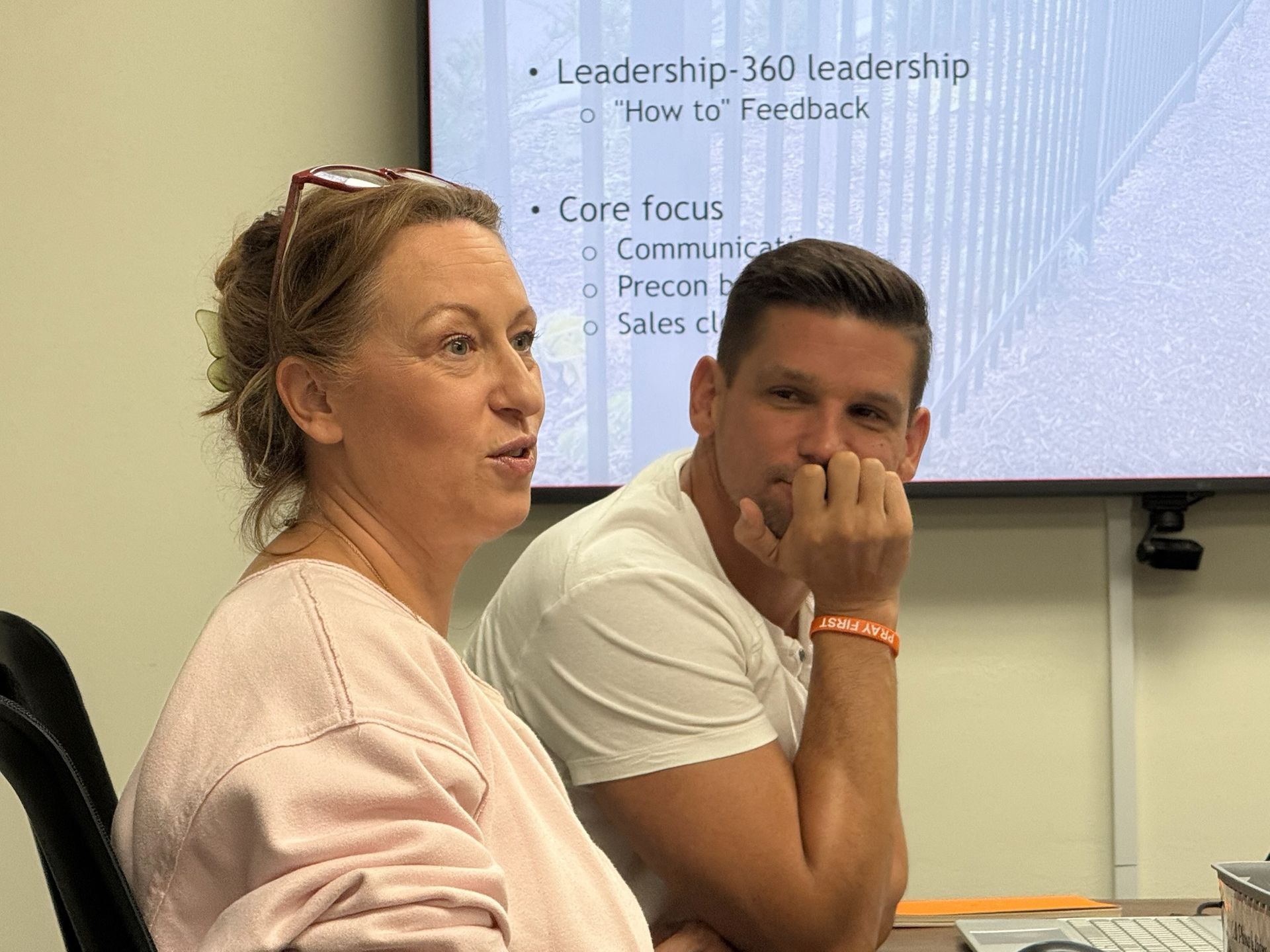 Woman speaking, man listening, both seated near a presentation screen with text.