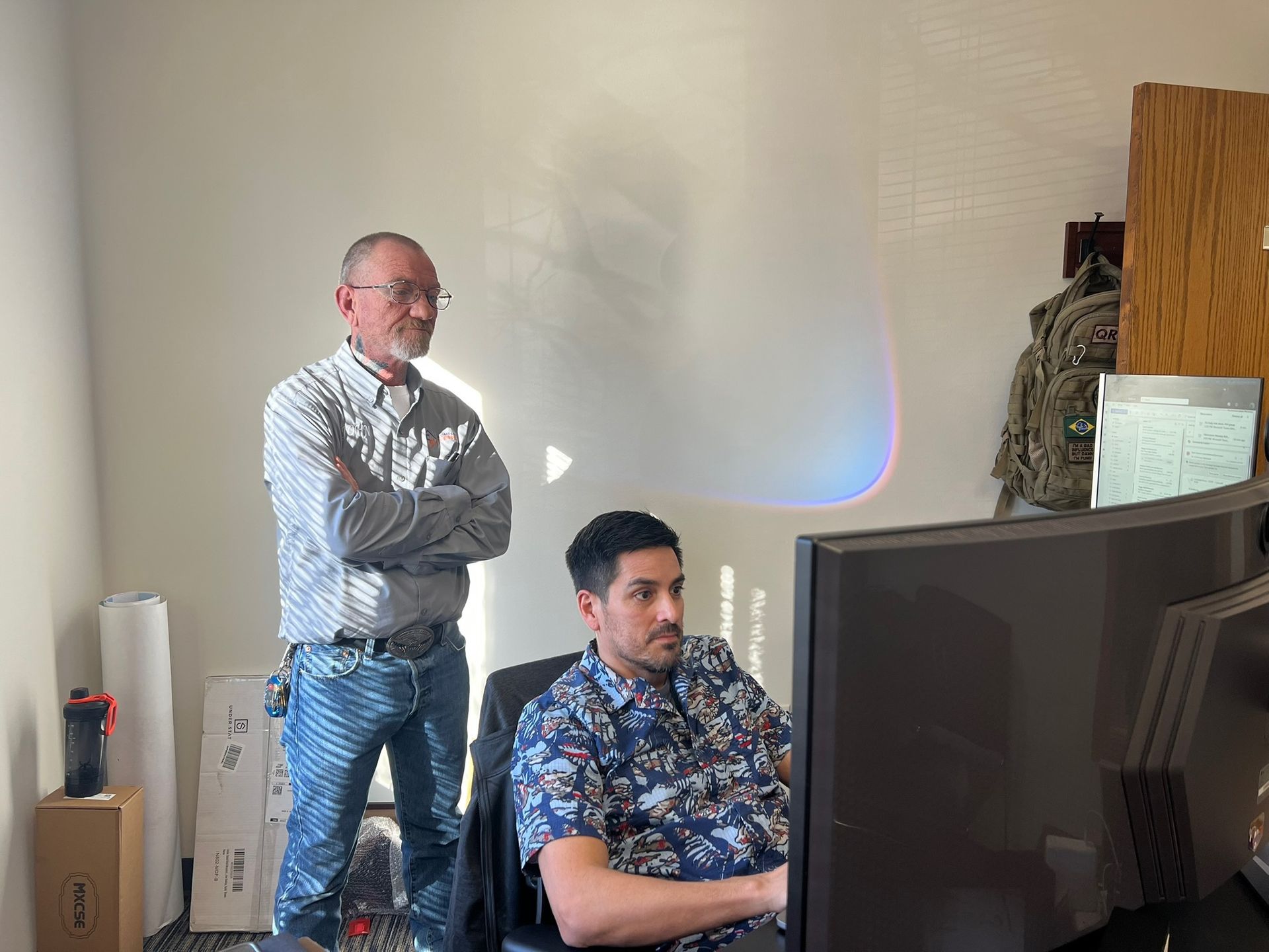 Man in patterned shirt seated at computer, man in striped shirt standing with arms crossed, in office.