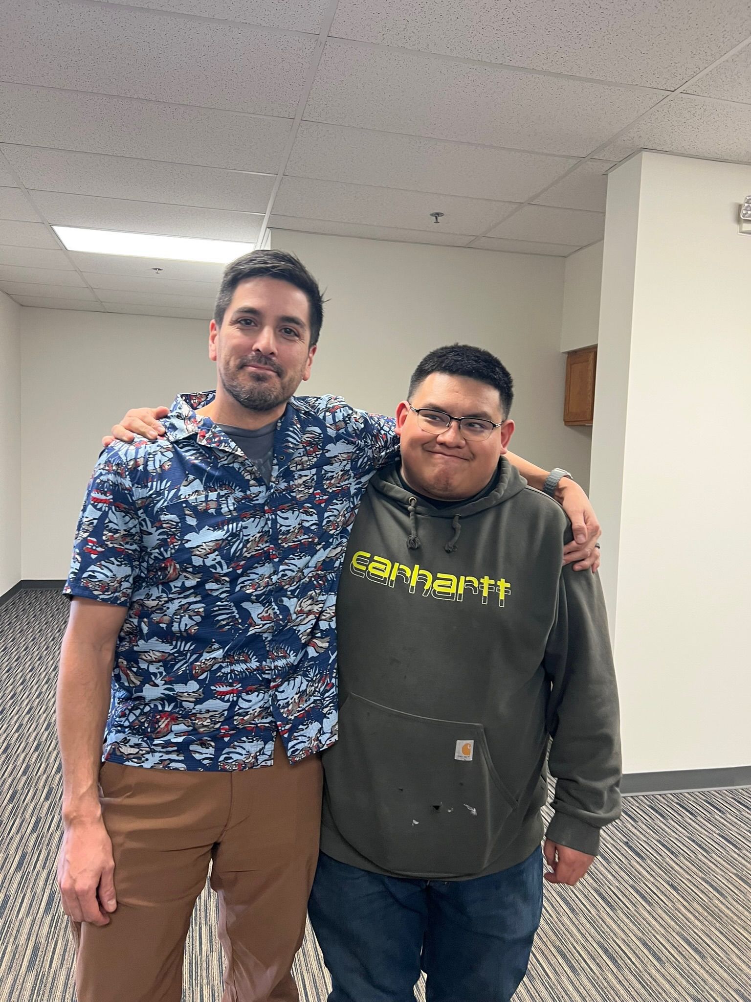 Two men with arms around each other posing for a photo. One wears a blue-patterned shirt and brown pants, the other a Carhartt hoodie.