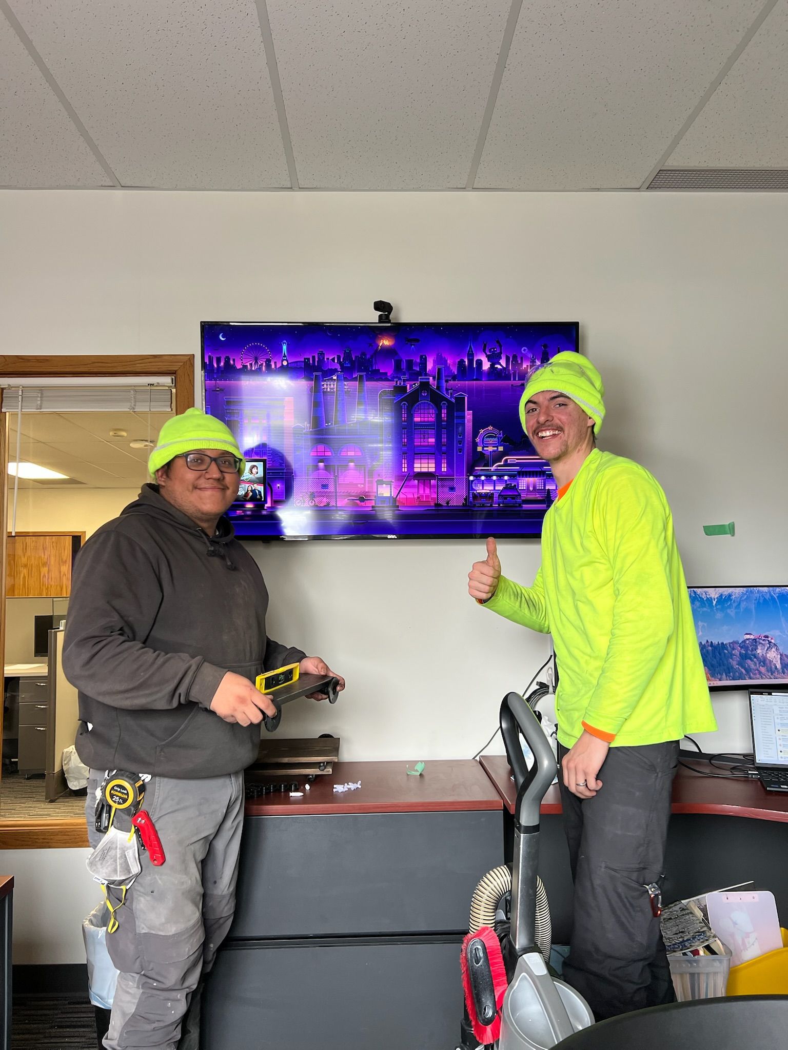 Two people in neon green hats and shirts stand by a TV. One holds tools, the other gives a thumbs up. Interior setting.