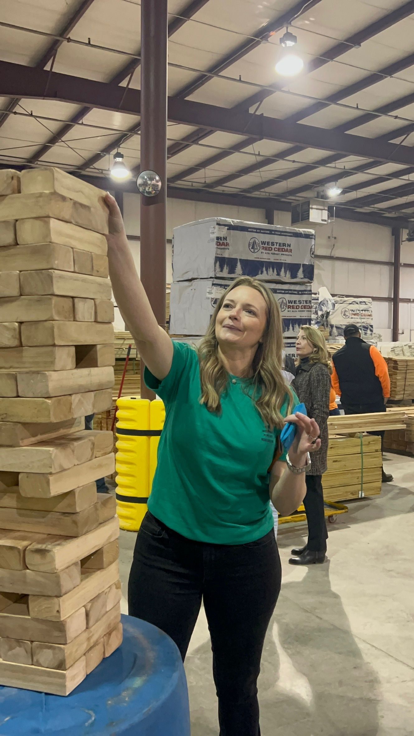 Woman in green shirt balances a ball on top of a giant Jenga tower in an industrial setting.