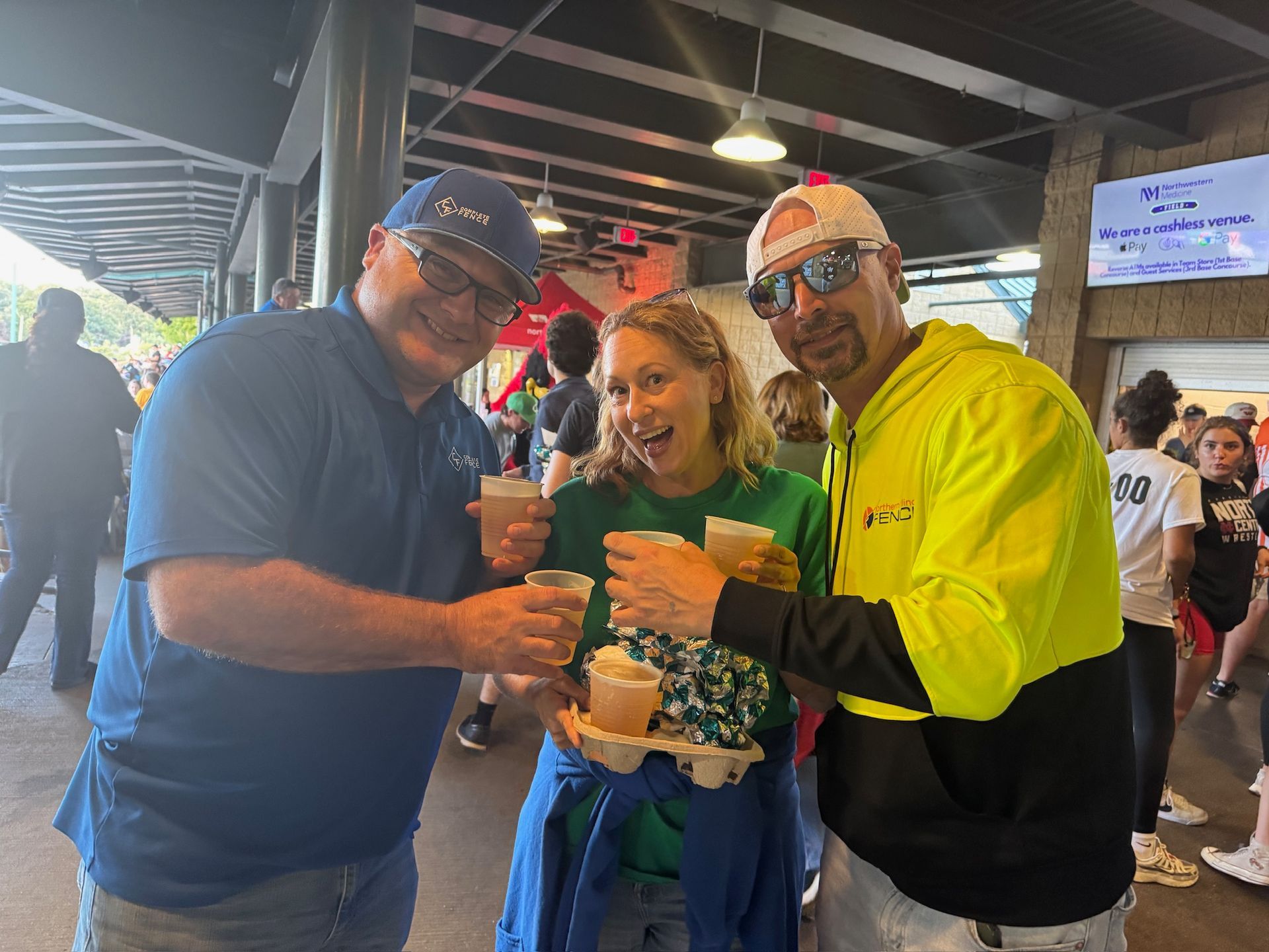 Three people holding drinks, smiling. Woman in green, men in blue and yellow. Outdoors, under awning.