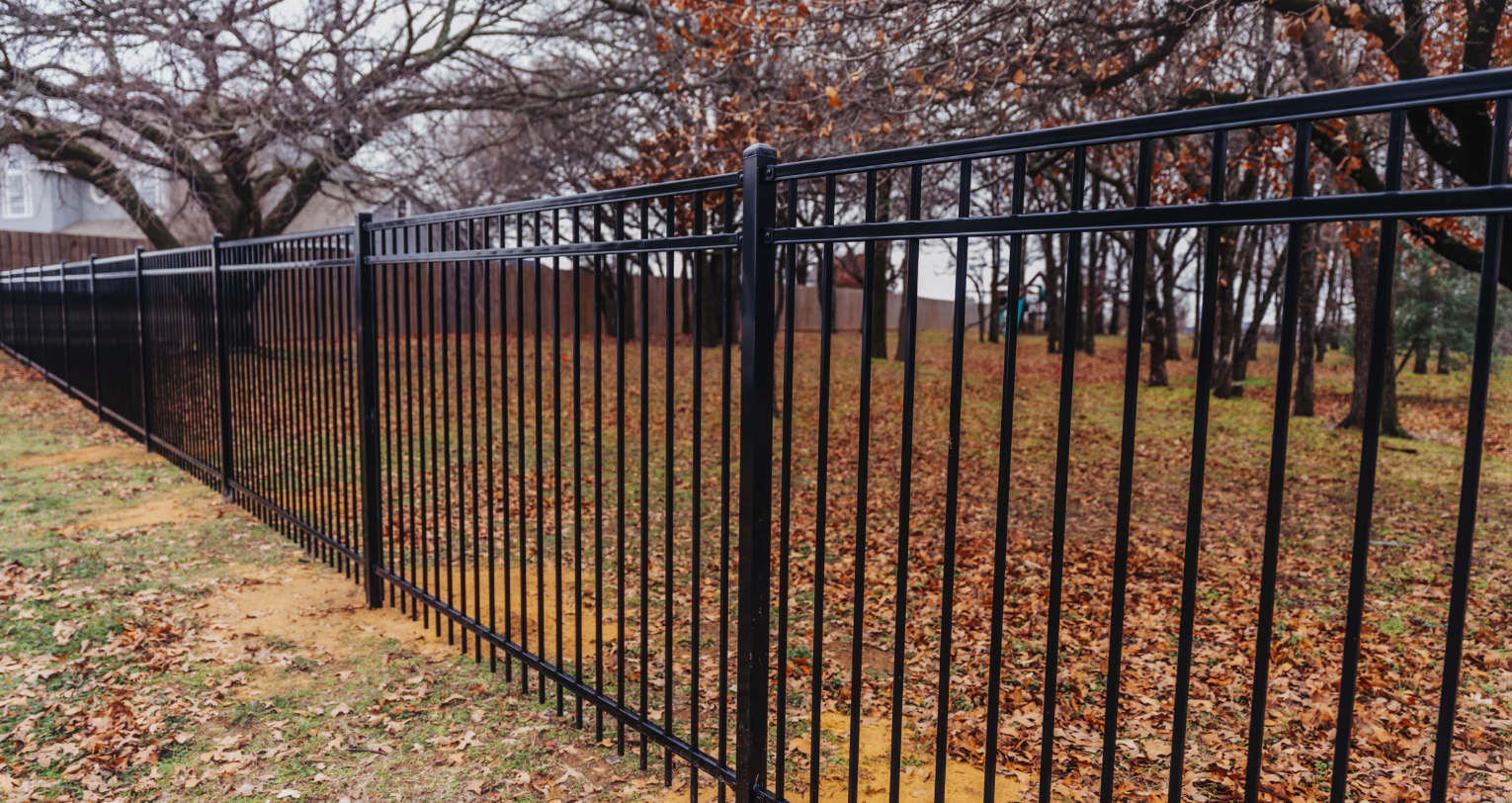 Black metal fence, outdoors. Brown leaves and trees in background.