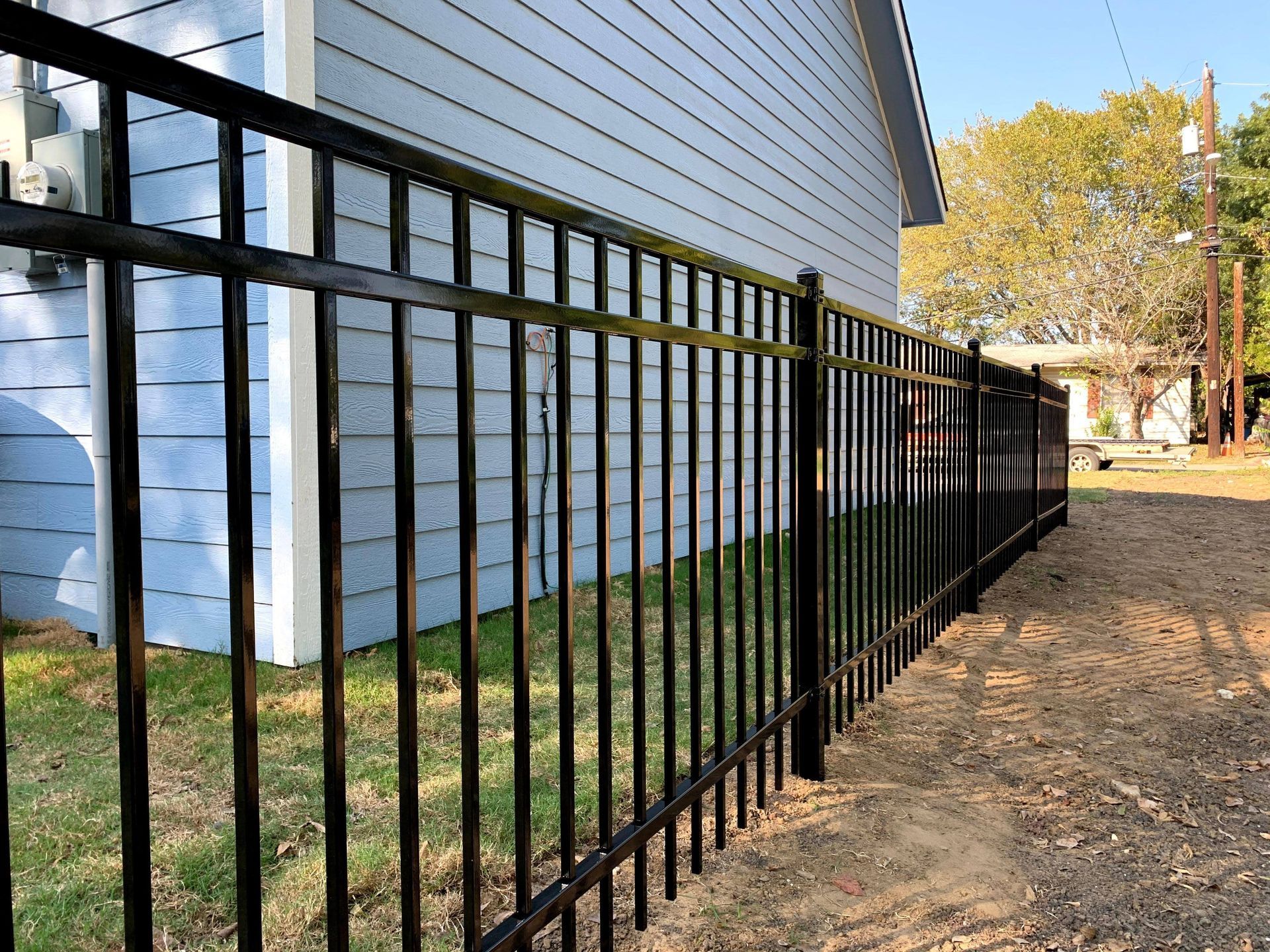 Black metal fence in front of a light blue building, with a grassy area and dirt path.