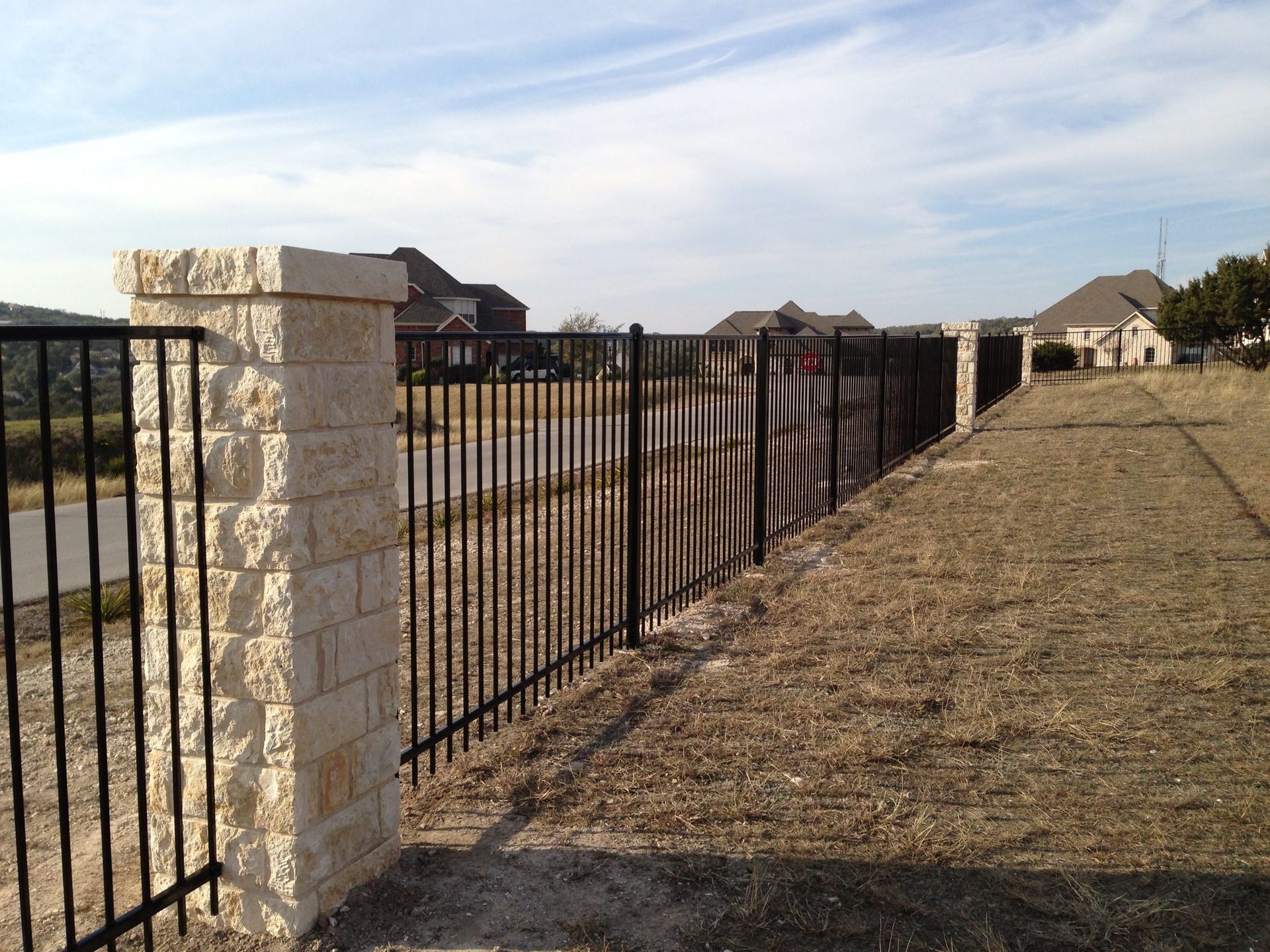 Black metal fence with stone pillars in a grassy area, houses in the background under a blue sky.