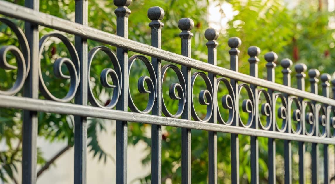 Black wrought iron fence with decorative swirls and ball finials, green foliage in background.