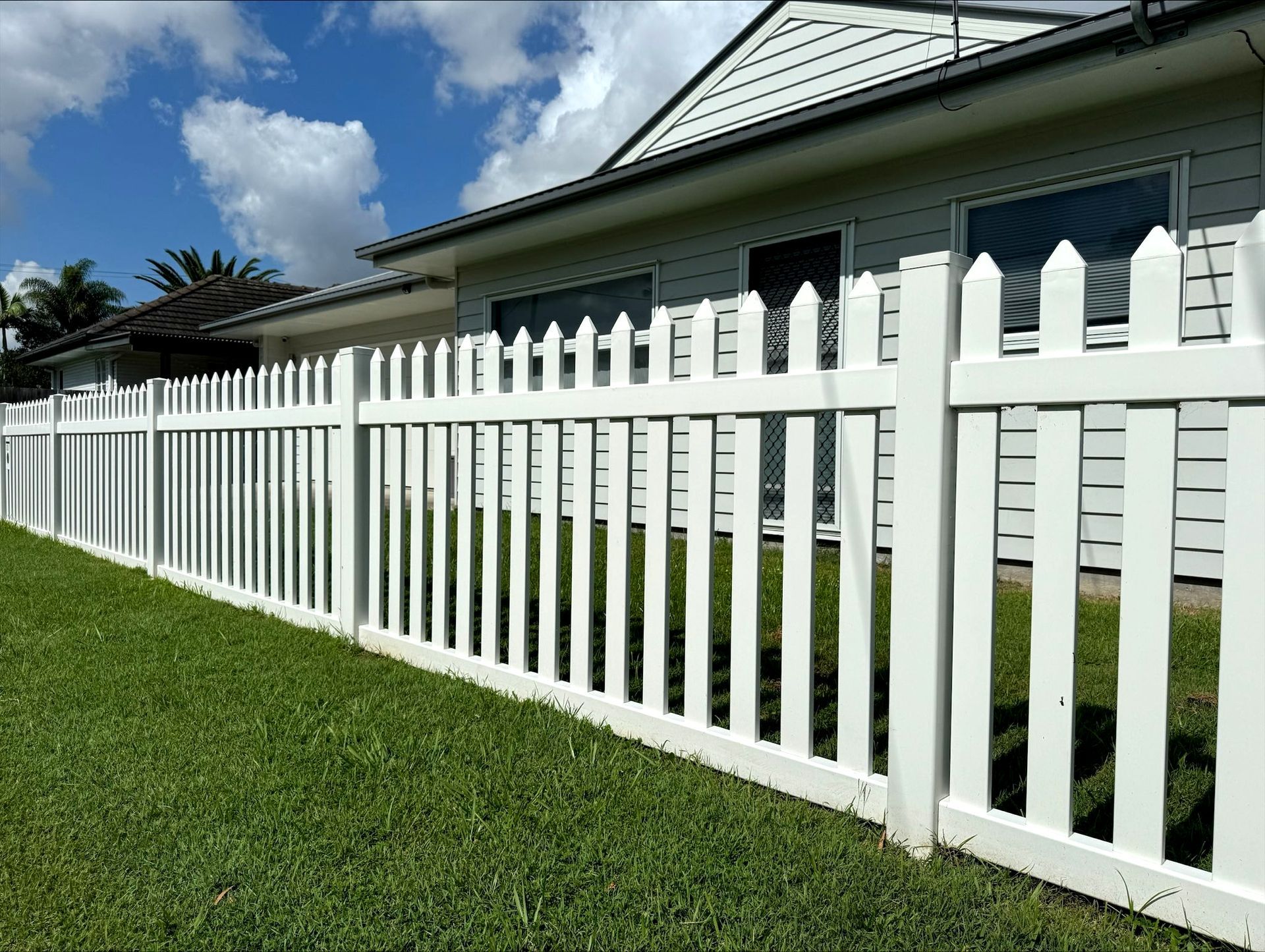 White picket fence in front of a white house with a green lawn under a blue sky.