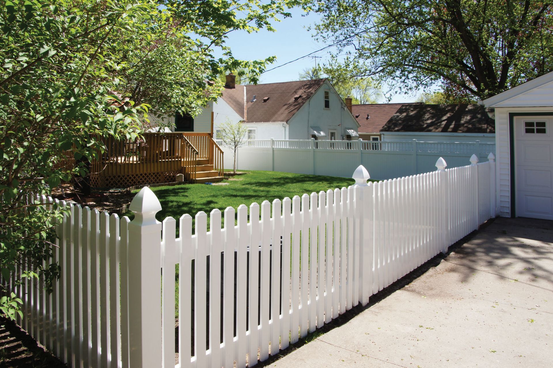White picket fence surrounding a backyard with a house, deck, and green lawn.